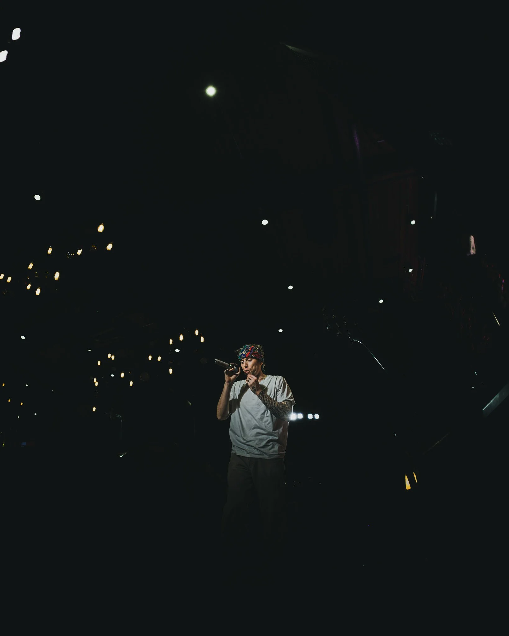 A person wearing a bandana and white t-shirt holding a microphone on stage with dark surroundings and small lights above.