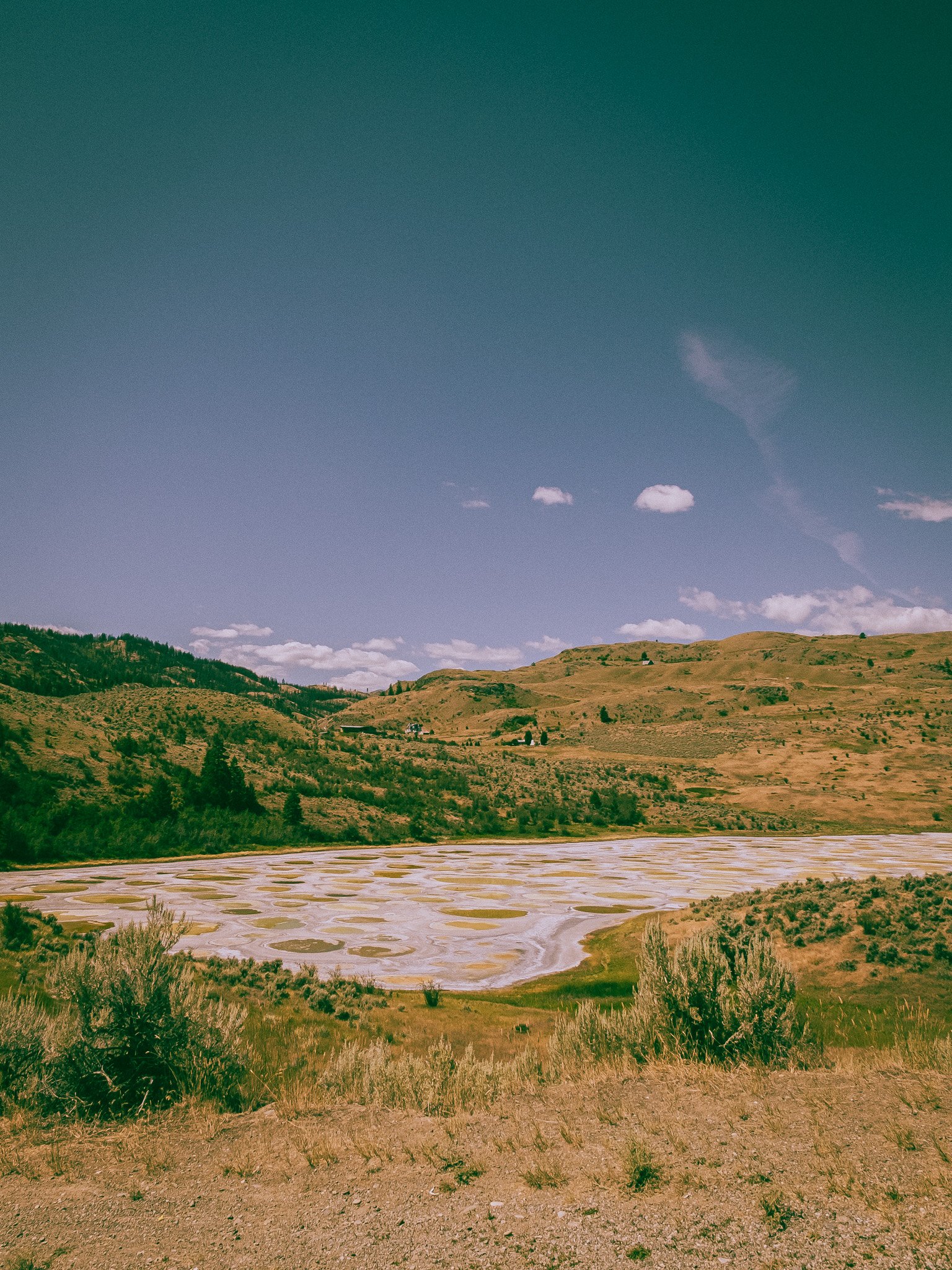 A landscape featuring a wide valley with brown, dry land, a salt flat with patterned white, light-colored mineral deposits, rolling grassy hills in the background, and a partly cloudy sky.