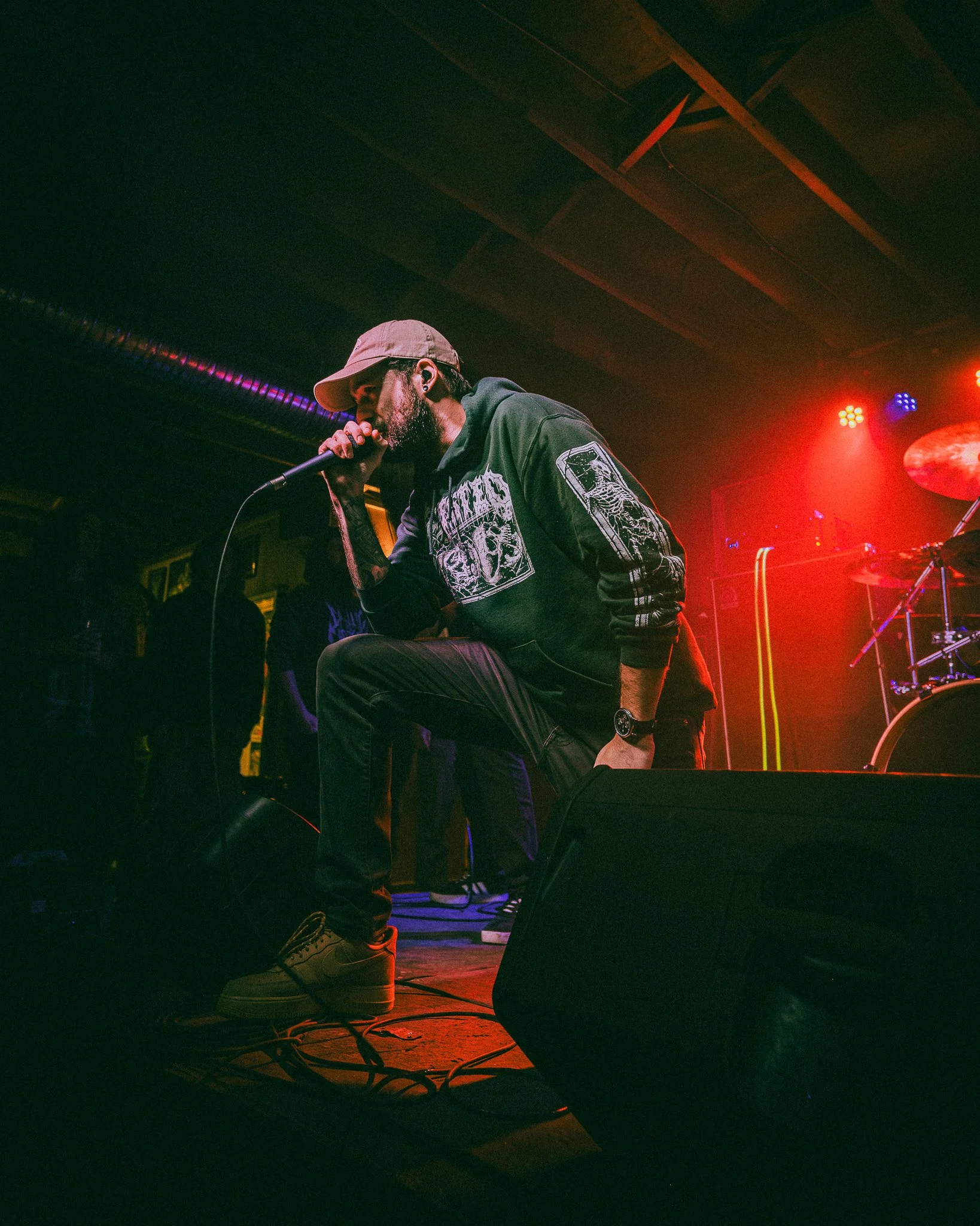 A male singer wearing a beige bucket hat, black hoodie with graphic designs, and black jeans, is performing on stage with a microphone, kneeling and singing passionately. The stage is bathed in red and purple lighting, with drums and other musical eq