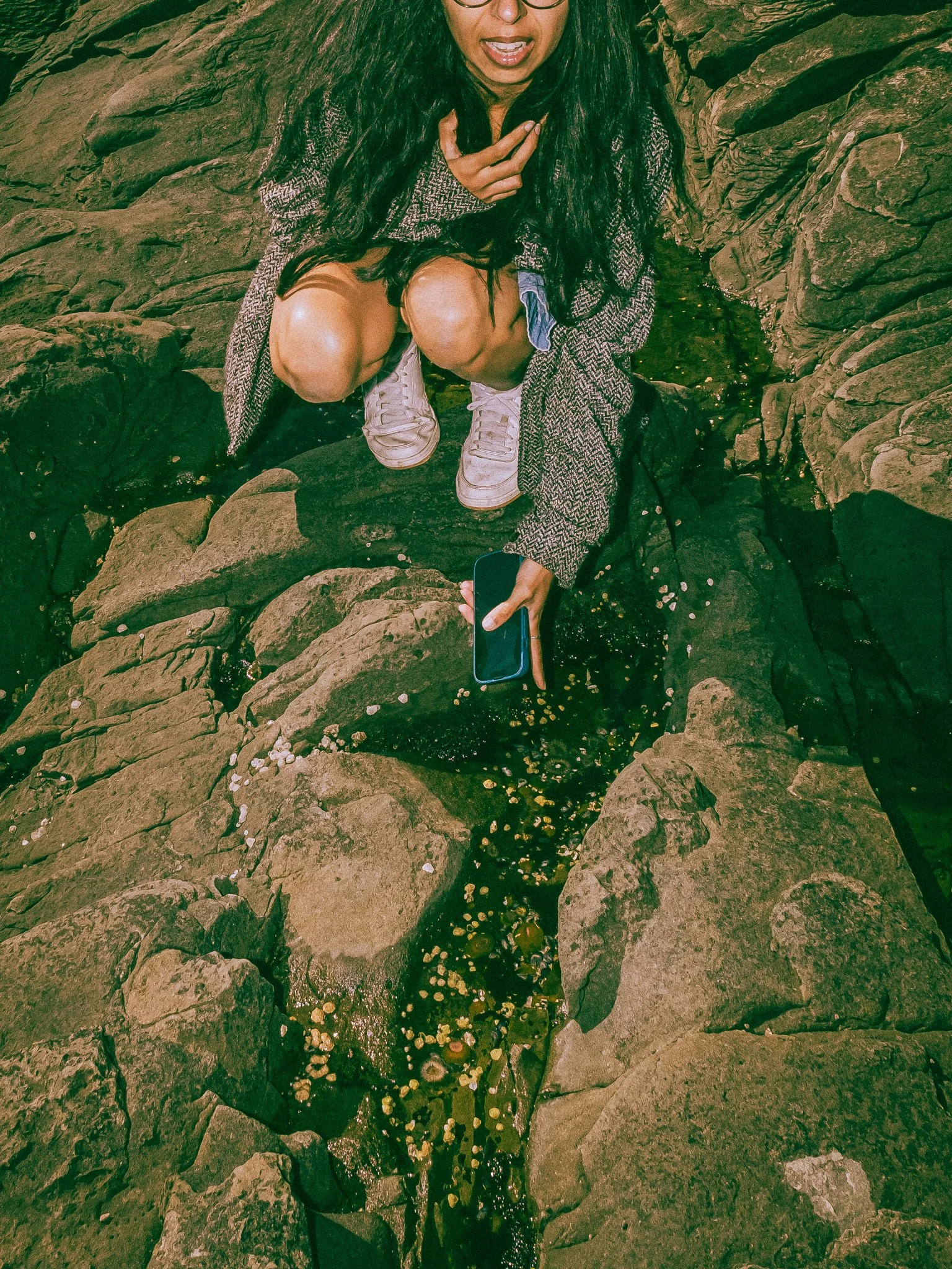 A woman with long dark hair and glasses crouches on rocks near water, holding a smartphone in her right hand, looking down, wearing a gray coat, white sneakers, and shorts.