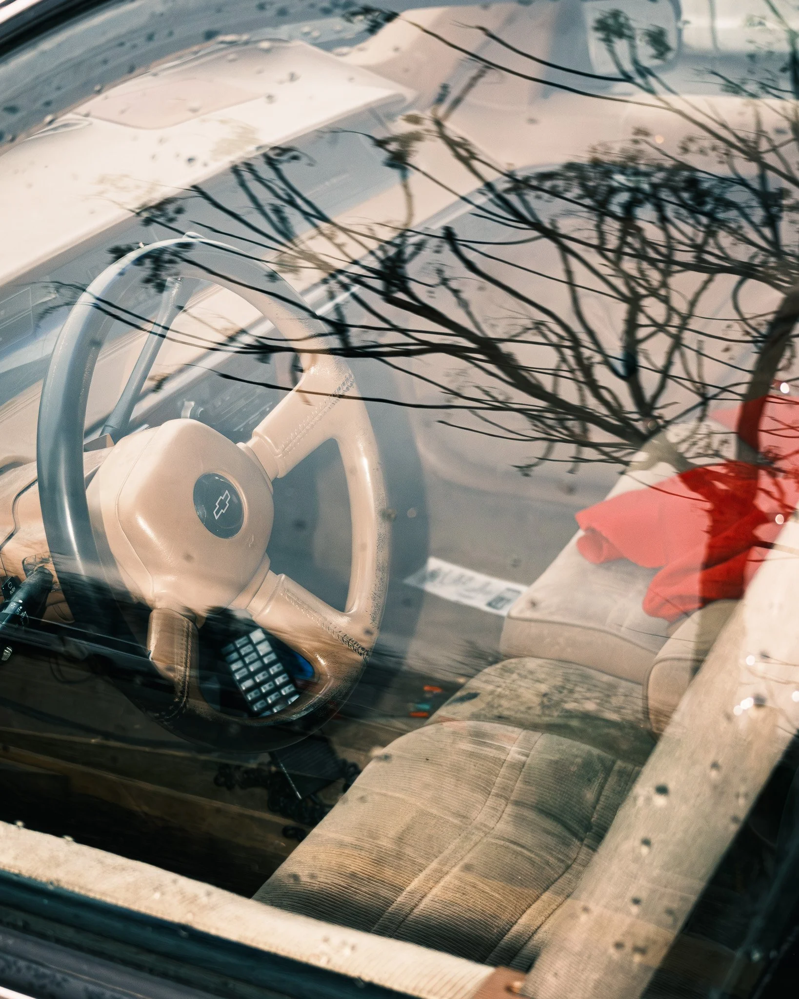 Interior of a vintage car with a Chevrolet emblem on the steering wheel, viewed through the windshield, showing the dashboard and front seat with a red piece of clothing or fabric on the seat.