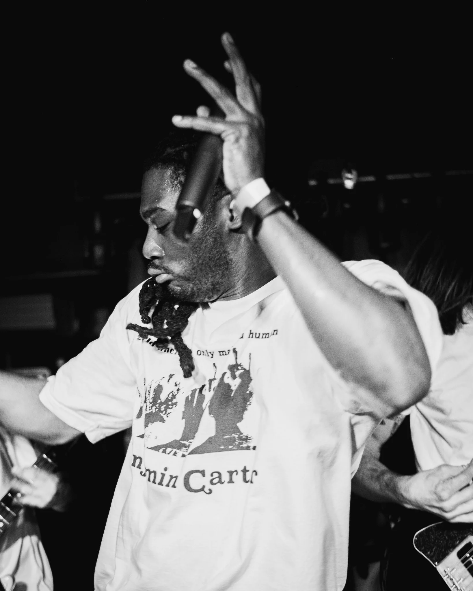 A young man with dreadlocks and dark skin is shown in profile, with his eyes closed, holding a microphone near his temple, wearing a T-shirt with a graphic and text, in a dark setting.