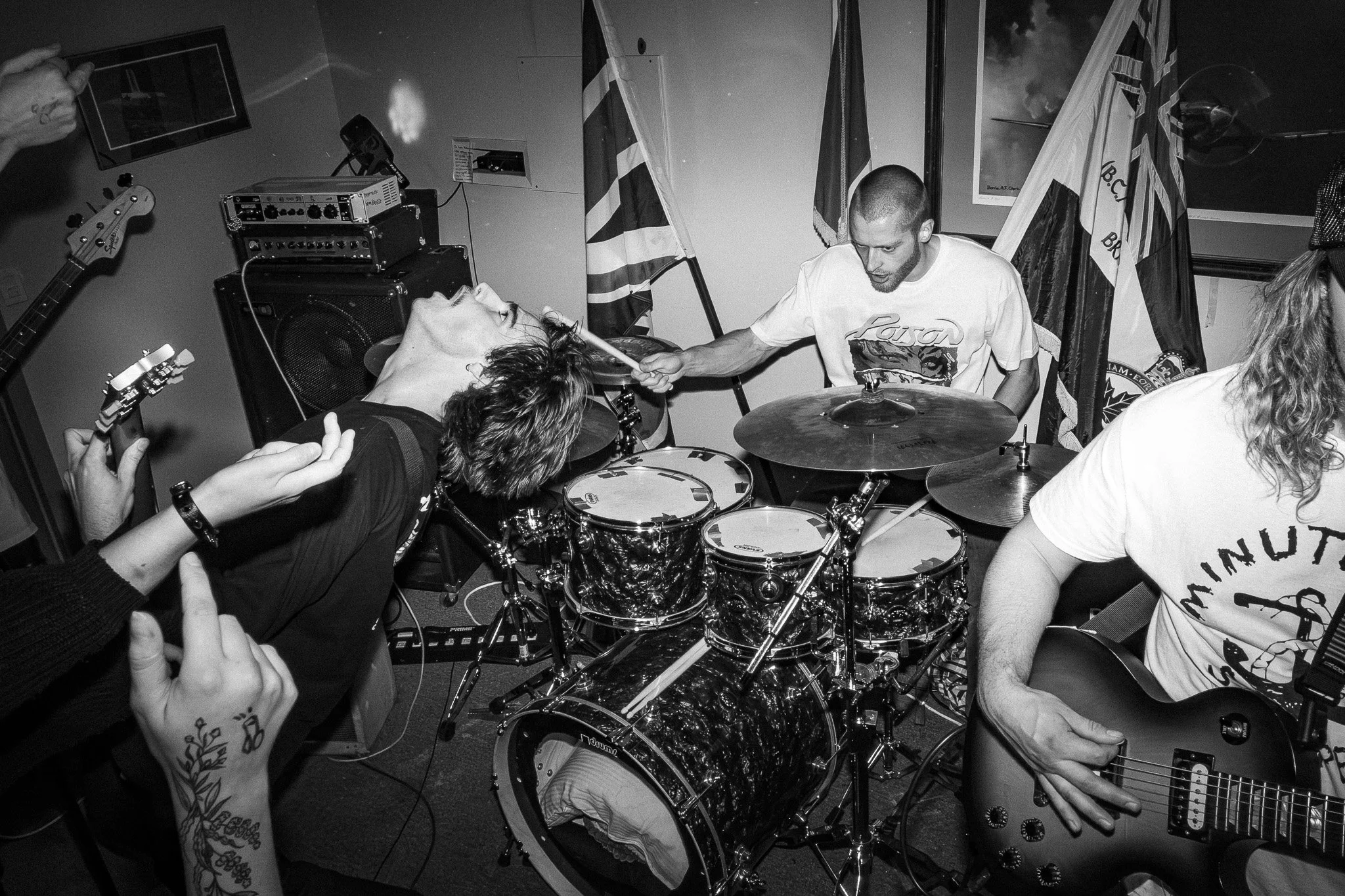 Black and white photo of a live music performance with a drummer, a guitarist, and other band members in a room decorated with flags.
