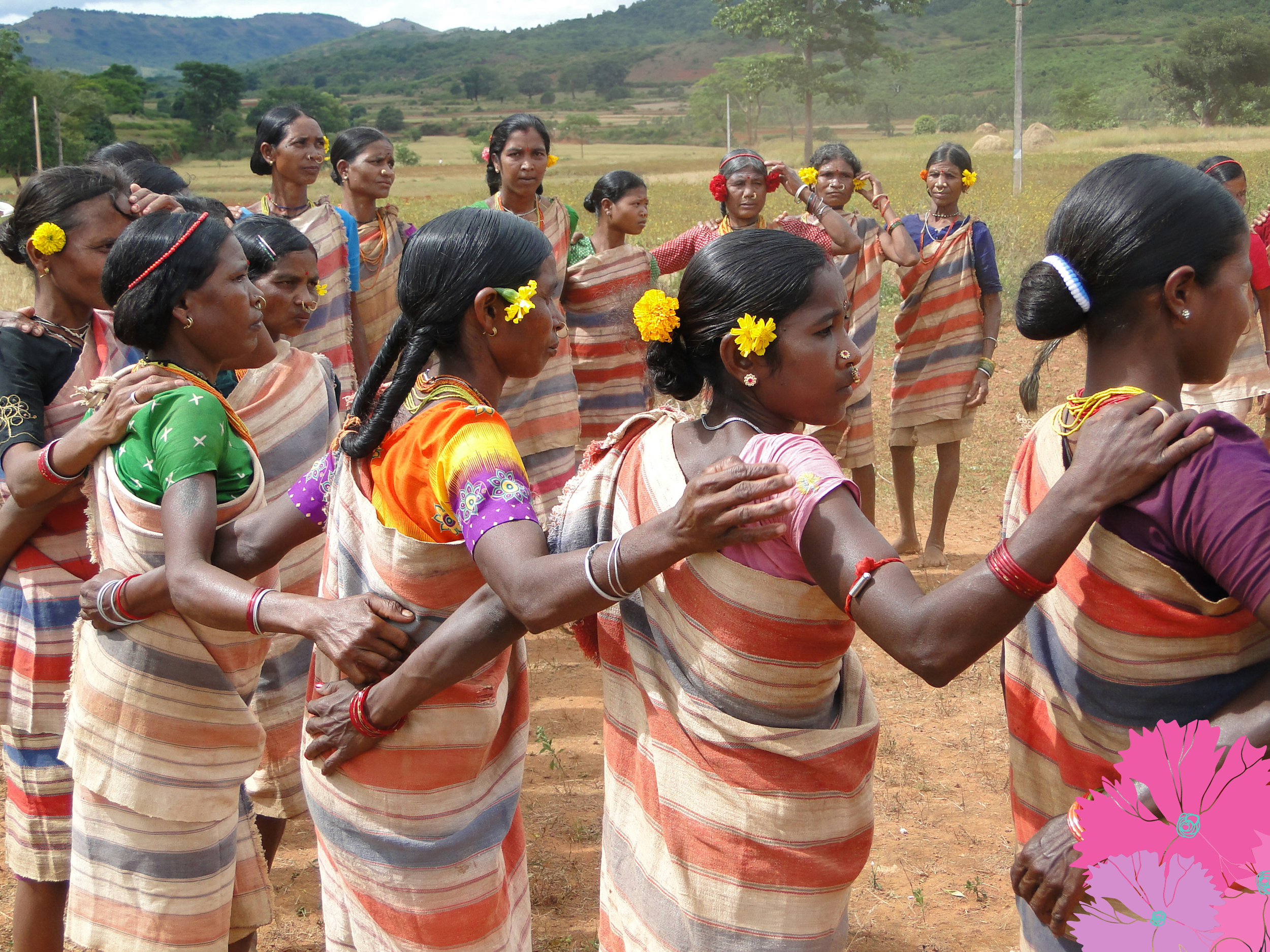 Group of Indian women dressed in traditional sarees, standing outdoors in a rural setting with hills and trees in the background, engaged in a dance or cultural activity.
