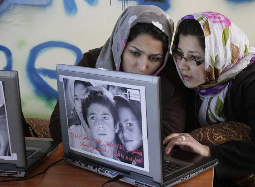 Two women wearing headscarves looking at a laptop screen displaying a black-and-white photo of children and Arabic text.