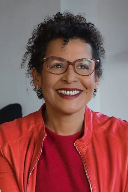 Woman with short curly hair and glasses wearing red leather jacket, smiling warmly.