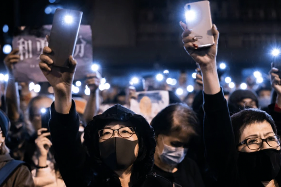 People gathered at a night-time protest or rally, holding up their phones with flashlight enabled, some wearing masks and glasses.