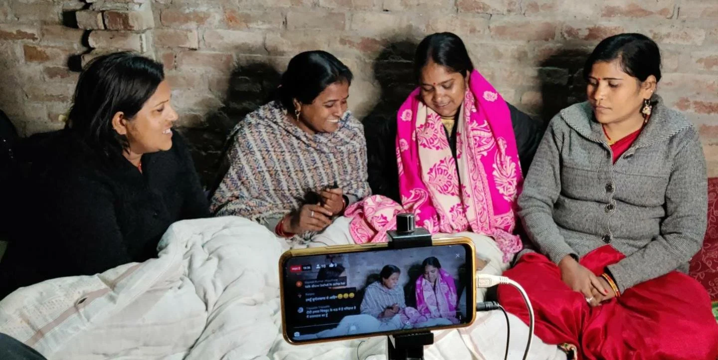 Four women sitting closely together in a room with brick walls, with a smartphone recording them facing each other.