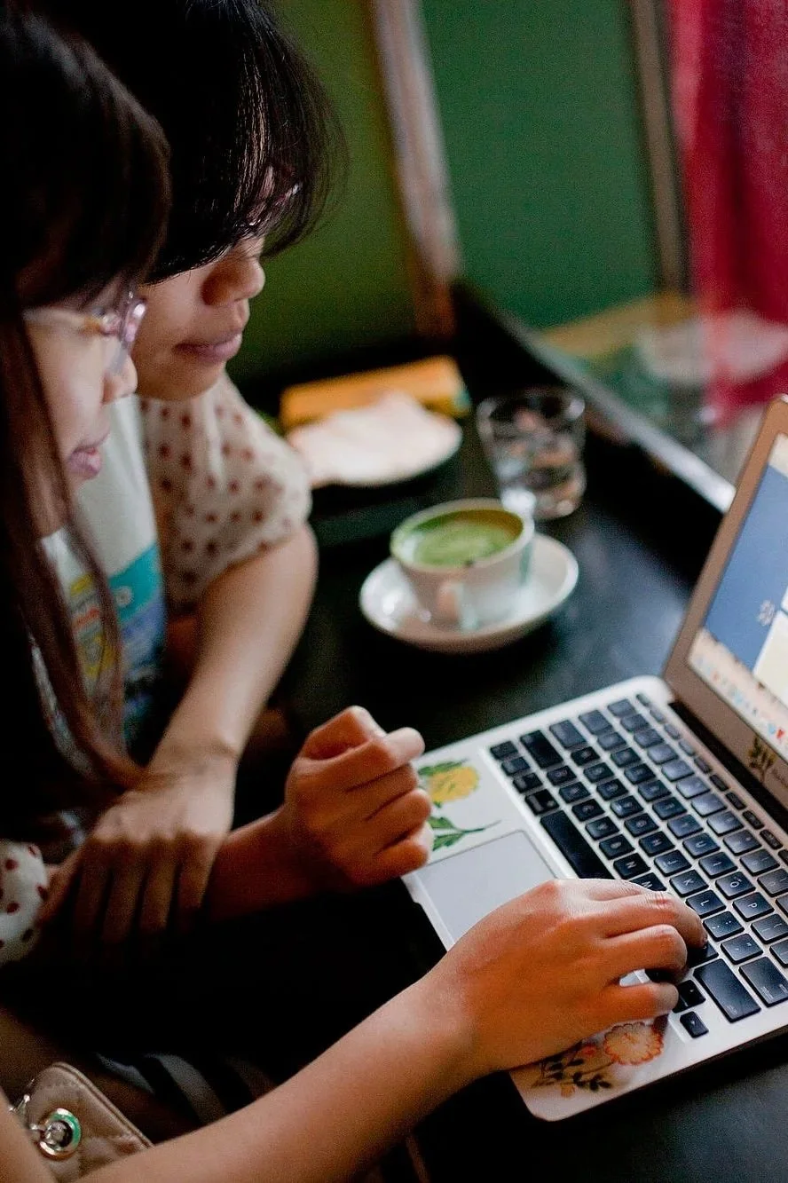Two young Vietnamese women are collaborating at a laptop in a cafe with a cup of green tea on the table.