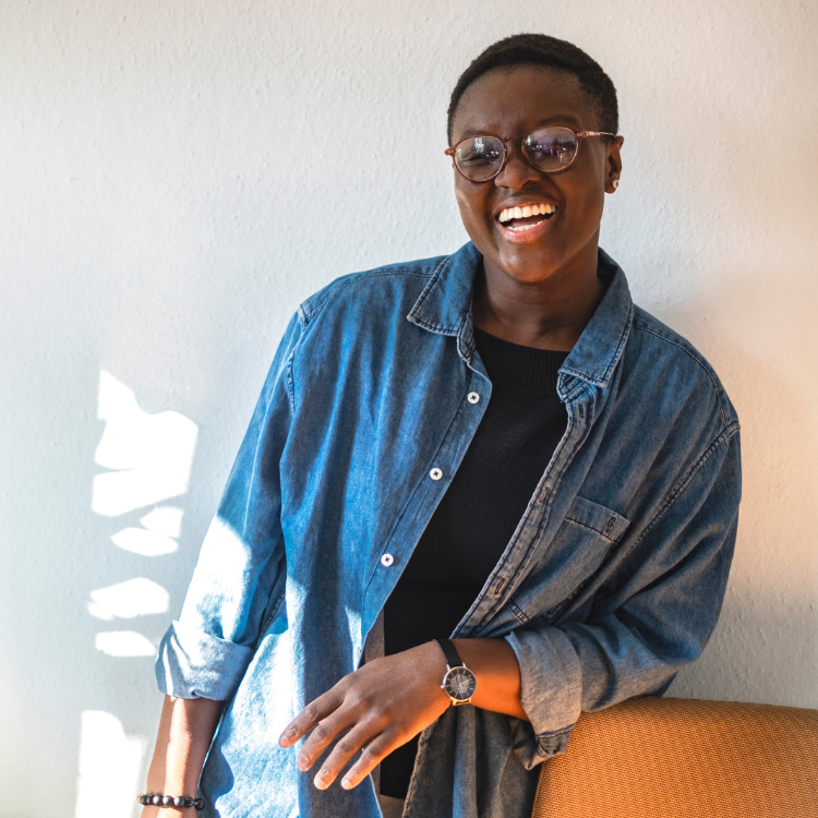 Black woman with short natural hair and glasses wearing denim jacket, smiling, smiling with a relaxed posture.