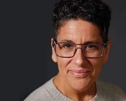 Person with short curly hair and glasses wearing gray, smiling