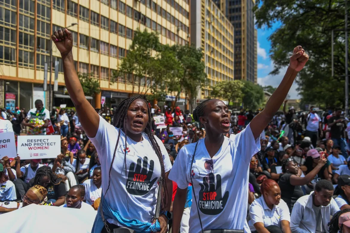 Women protesting against femicide, holding signs and wearing shirts that say 'Stop Femicide' in a city street with a crowd and tall buildings.