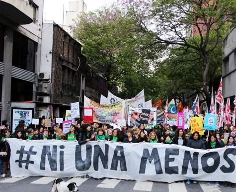 A large group of people protesting on a city street, holding a banner that says '#NiUnaMenos' with numerous signs and flags among the crowd.