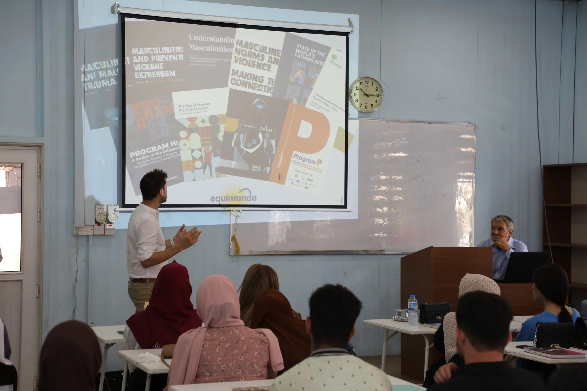 A classroom lecture scene with a male presenter standing in front of a large screen displaying multiple magazine covers or posters related to masculinity, violence, and social programs. Several students are seated at desks, watching the presentation, with some taking notes. A clock shows around 2:07, and a man is seated at a desk on the right, observing the presentation.