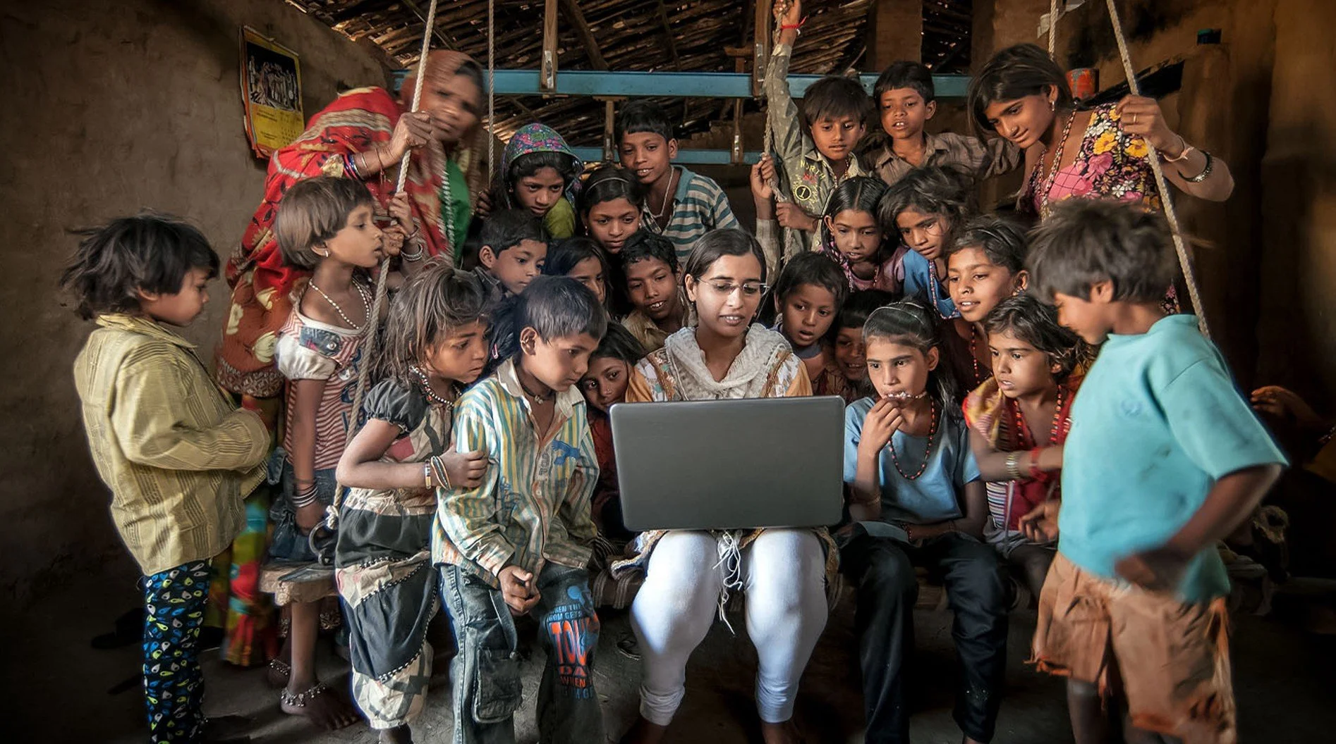 A woman sits with a group of children gathered around her, looking at a laptop screen inside a rustic room.