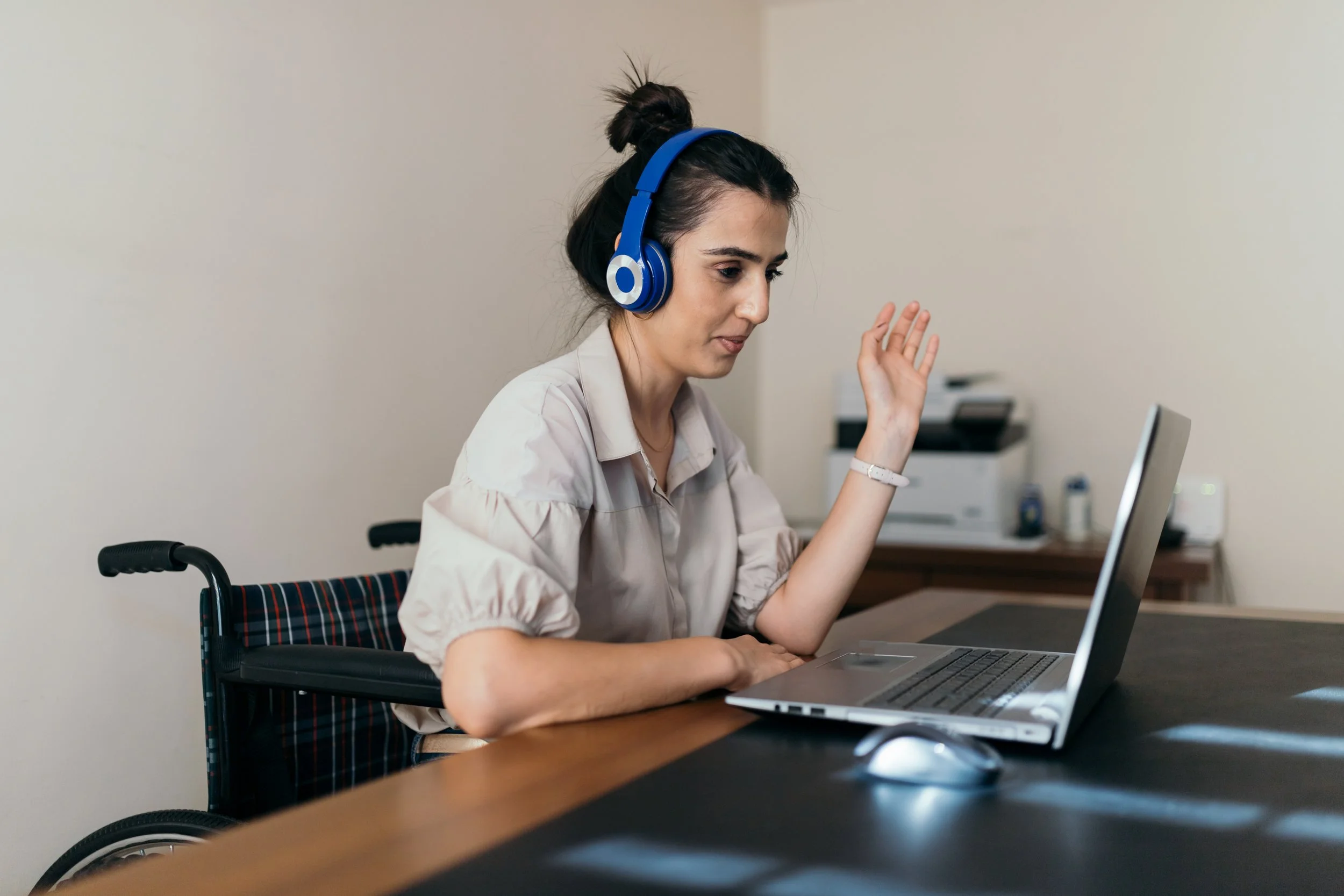 Woman in a wheelchair wearing headphones, sitting at a desk and engaging in a video call on her laptop.