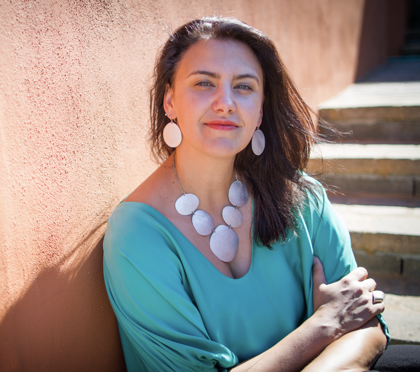 White woman with brown wavy hair wearing turquoise and silver jewelry, photographed outdoors against an orange wall with sunlight shining on her face.