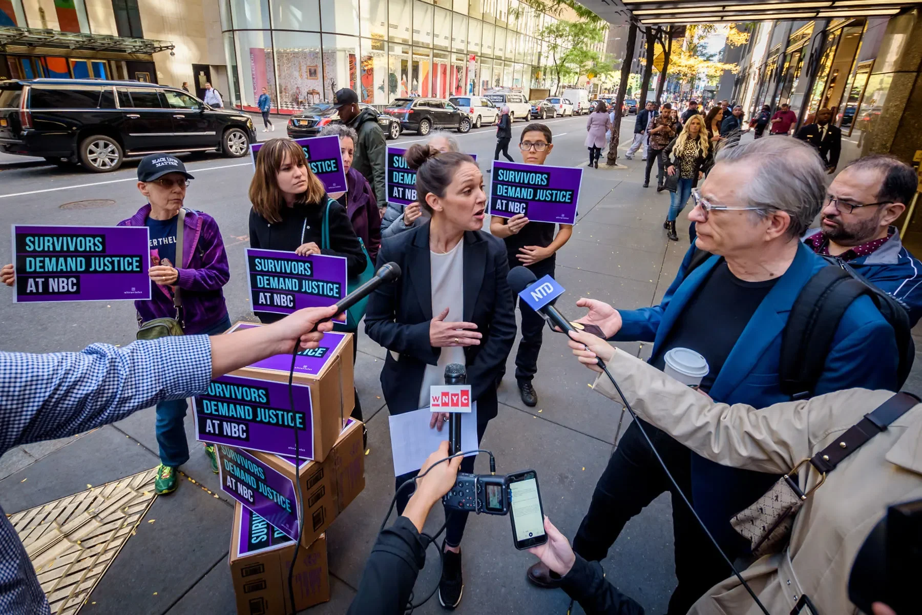 A group of protesters holding purple signs that read "Survivors Demand Justice at NBC" standing on a city sidewalk while being interviewed by multiple news reporters and journalists.