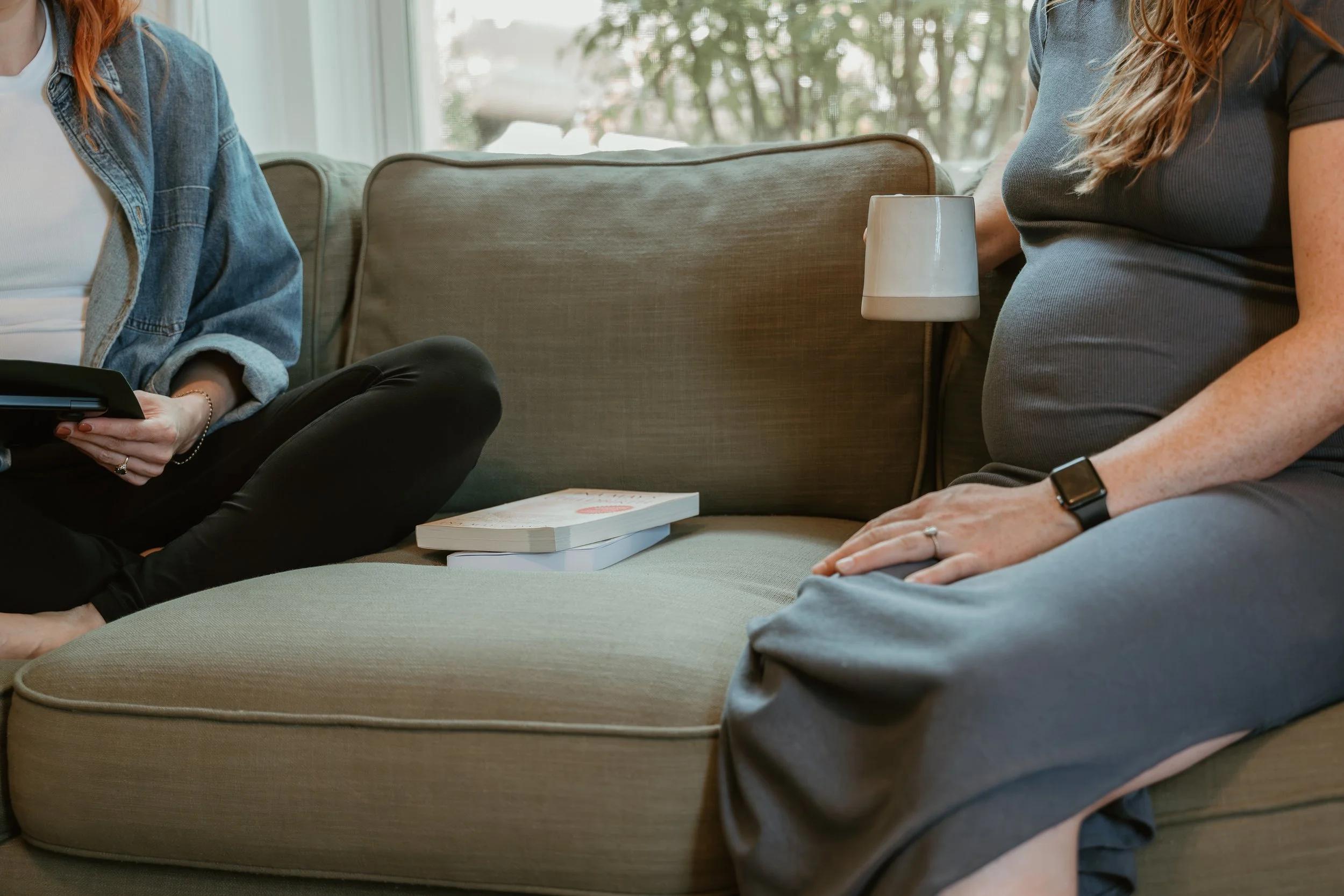 A pregnant woman sitting on a green sofa with a book and a lamp nearby, engaged in a conversation with another woman holding a tablet, in a bright living room.