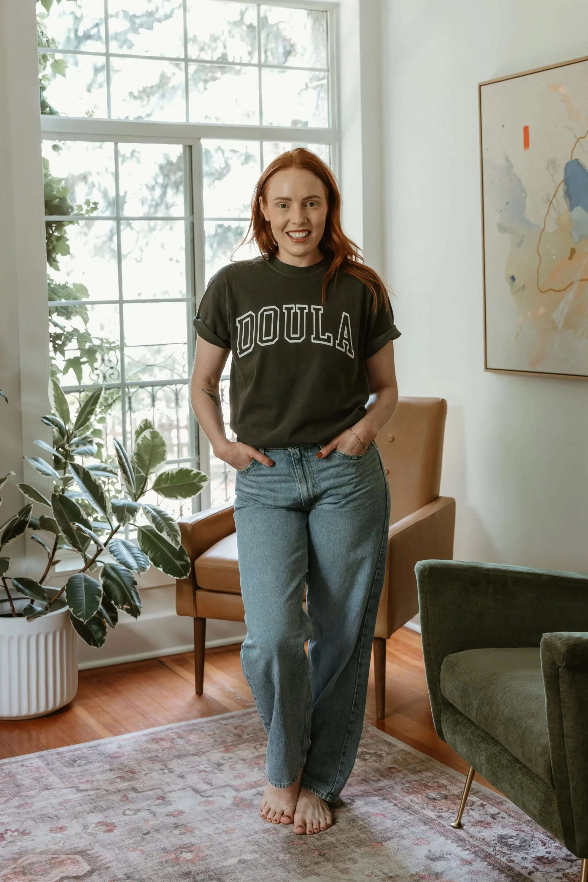 A woman with red hair, wearing a black t-shirt with 'DOULA' printed on it and blue jeans, standing barefoot in a living room with large window, plants, and modern décor.