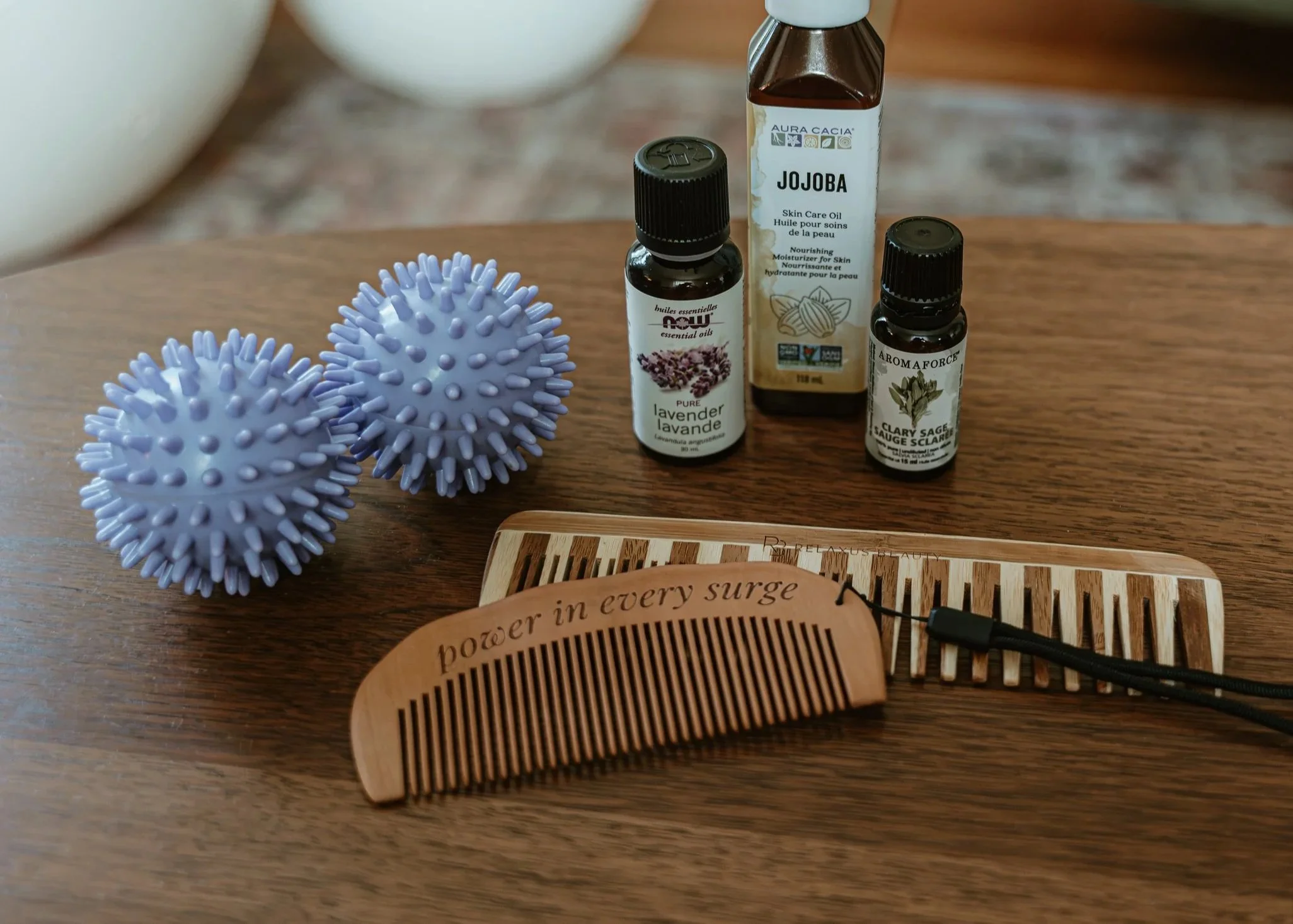 Two purple textured massage balls, bottles of essential oils labeled lavender, jojoba, and sage, a wooden comb with the inscription 'power in every surge', and a wooden hairbrush on a wooden surface.
