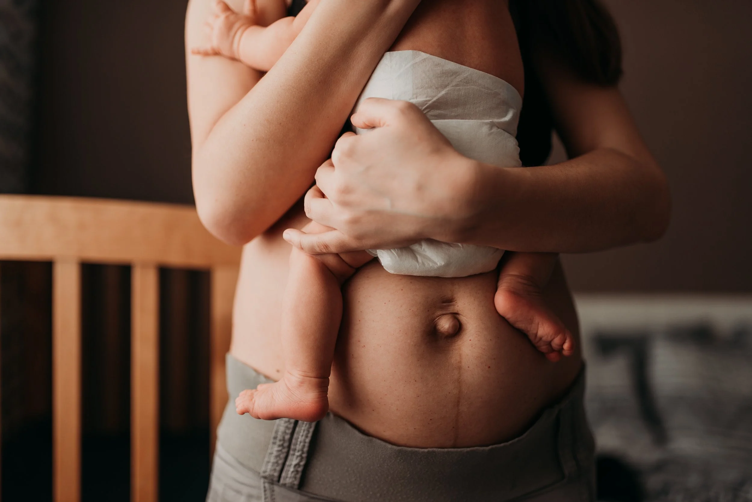 A person is holding a small baby against their chest. The adult's arms are wrapped protectively around the baby, who is wearing a diaper. The adult's bare midsection and the baby's tiny foot are visible.