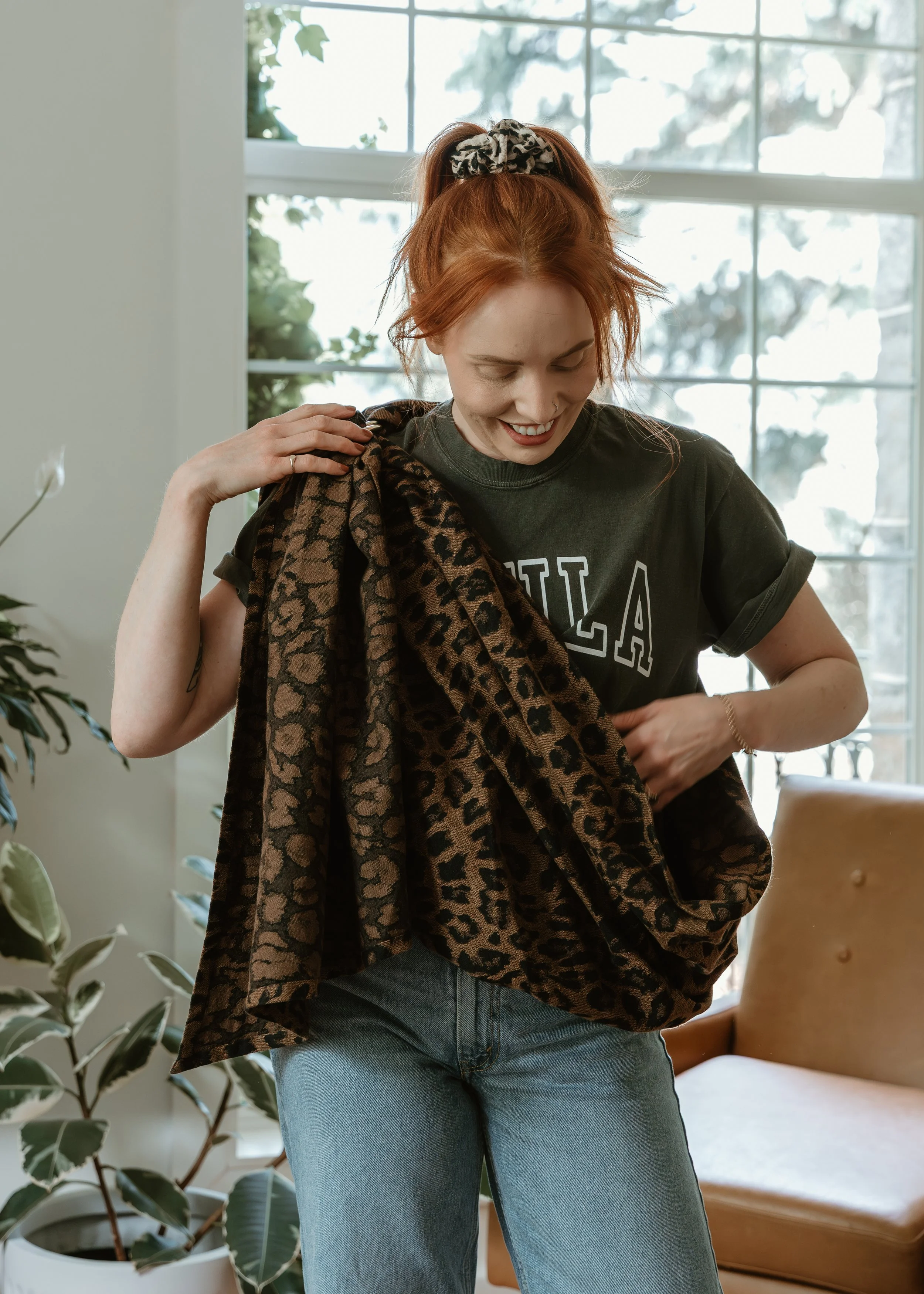 A woman with red hair tied up with a leopard print scrunchie is smiling and looking down while adjusting her baby carrier ring sling a bright room with large windows and plants.