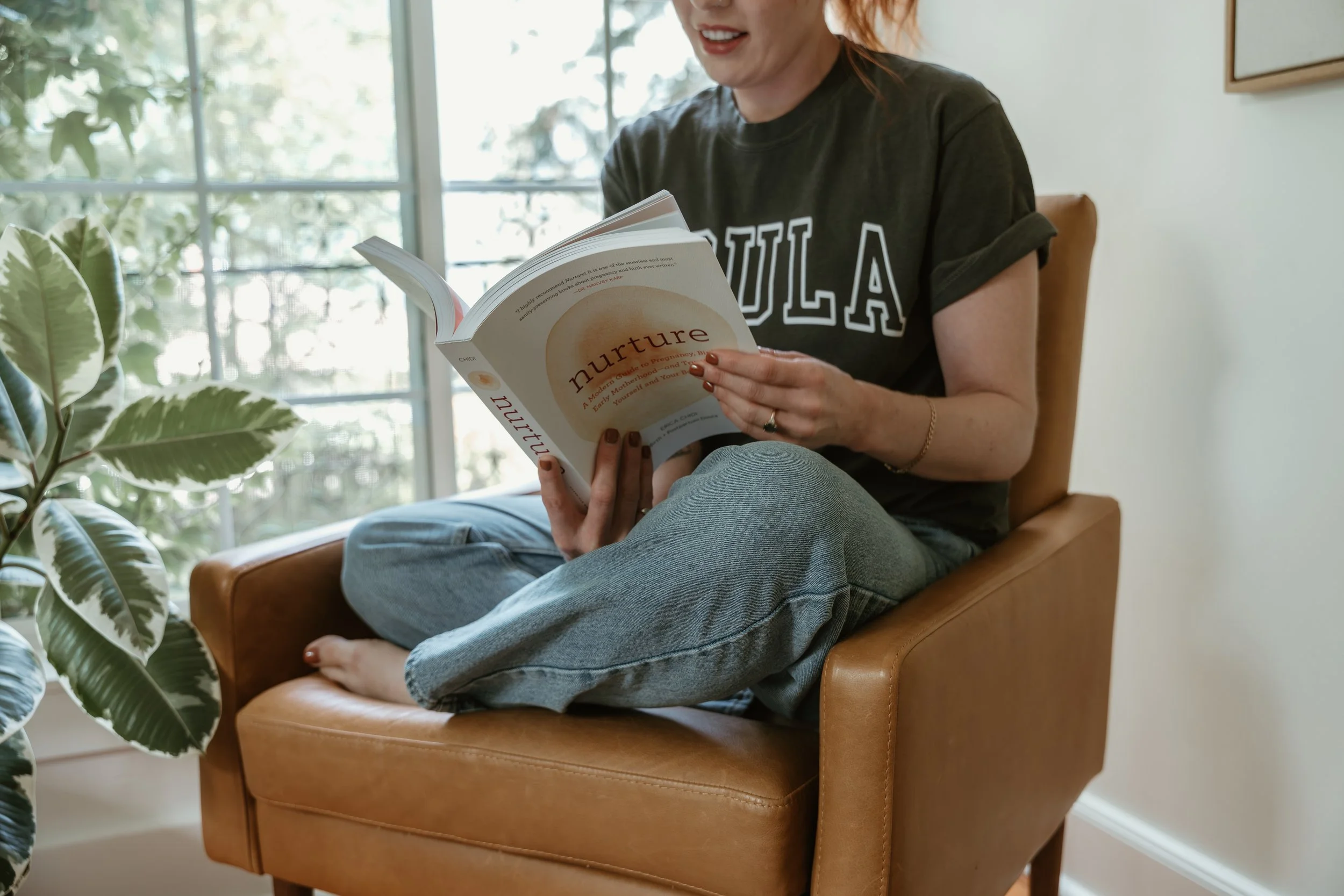 Woman sitting cross-legged on a tan leather chair, reading a book titled 'Nurture,' near a large window with green plants outside.