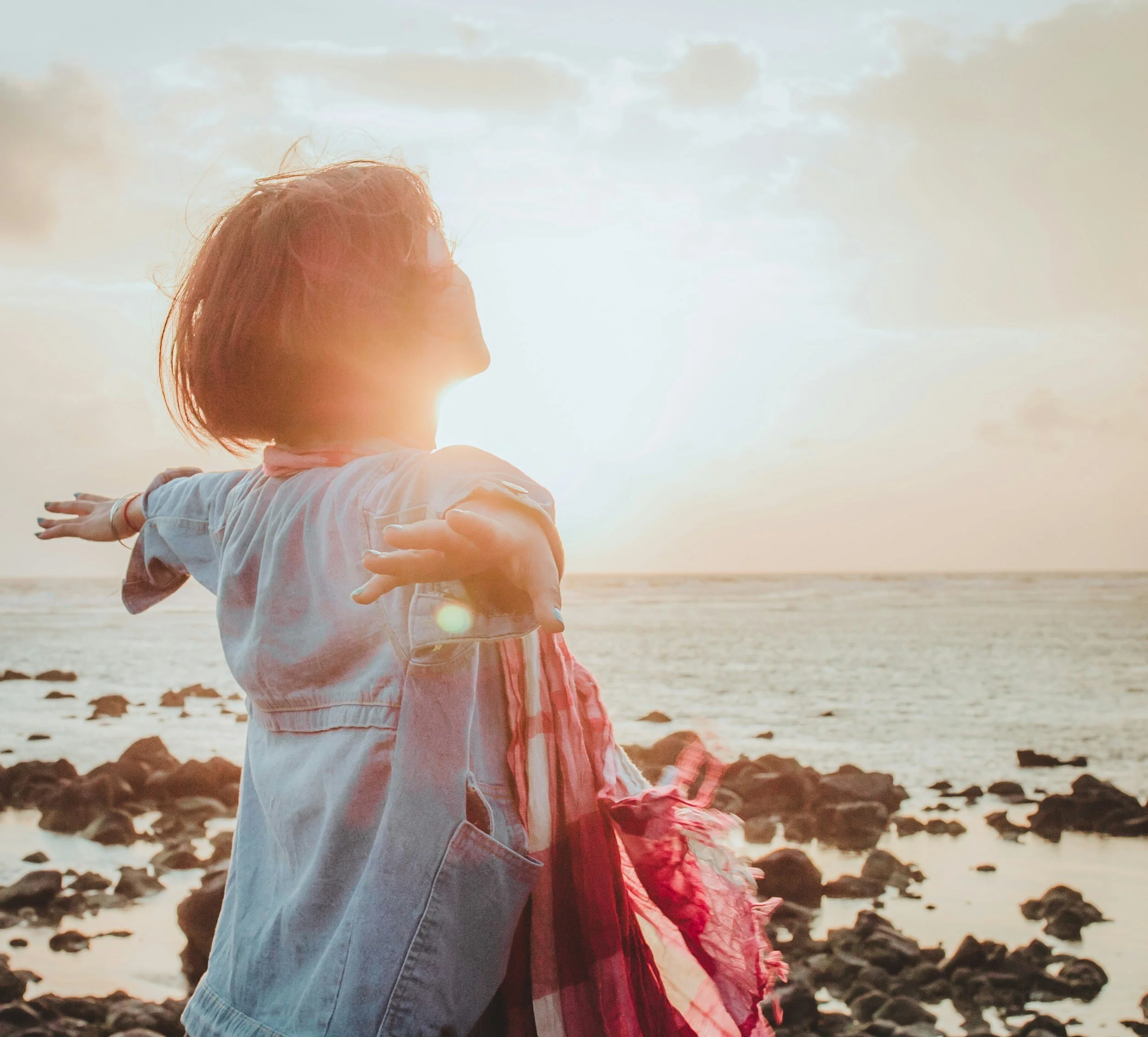 A woman with short hair and a light denim jacket standing on a rocky beach at sunset with her arms outstretched and a scarf in her hand.