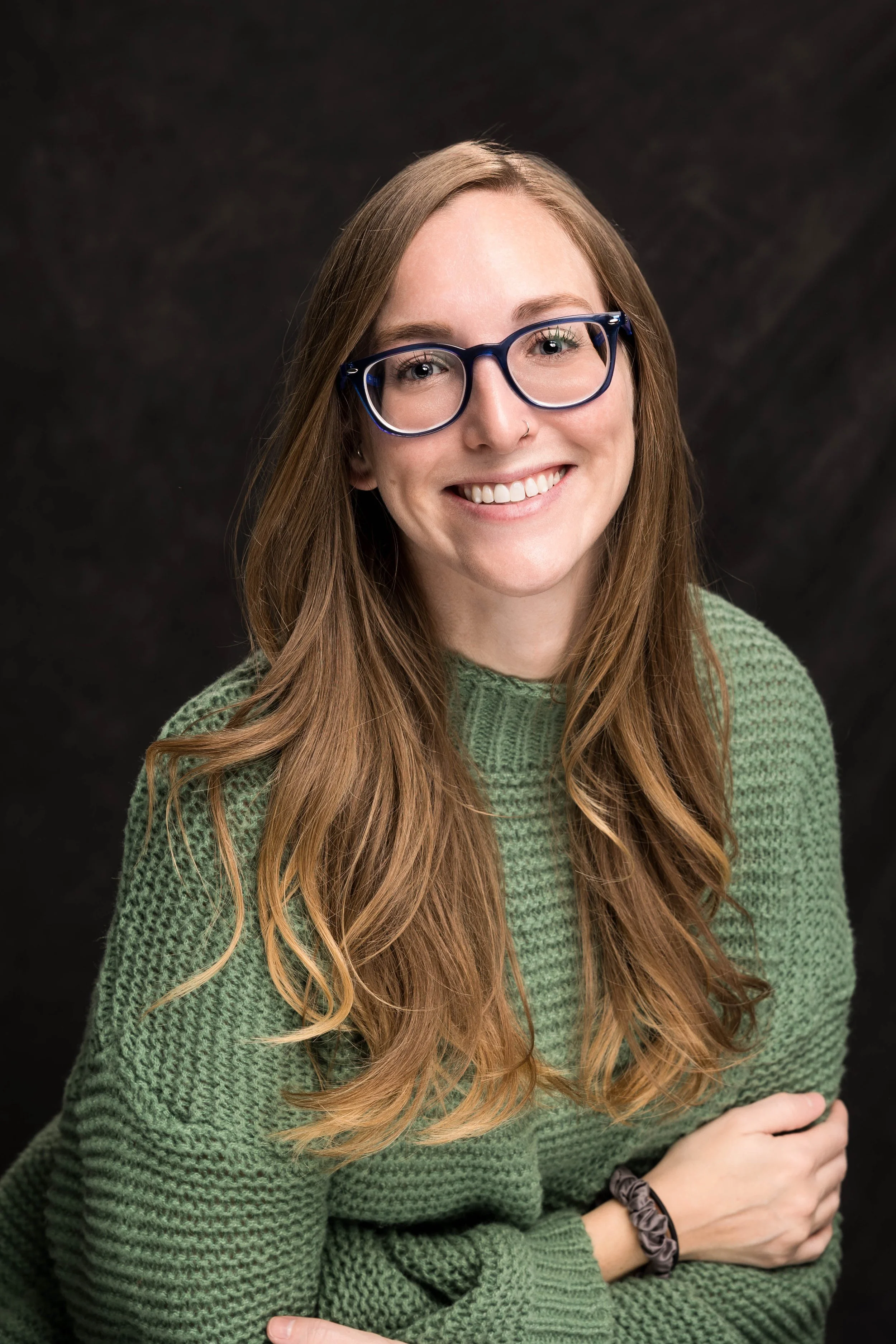 Young woman with long red hair, wearing blue glasses and a green knitted sweater, smiling at the camera against a dark background.