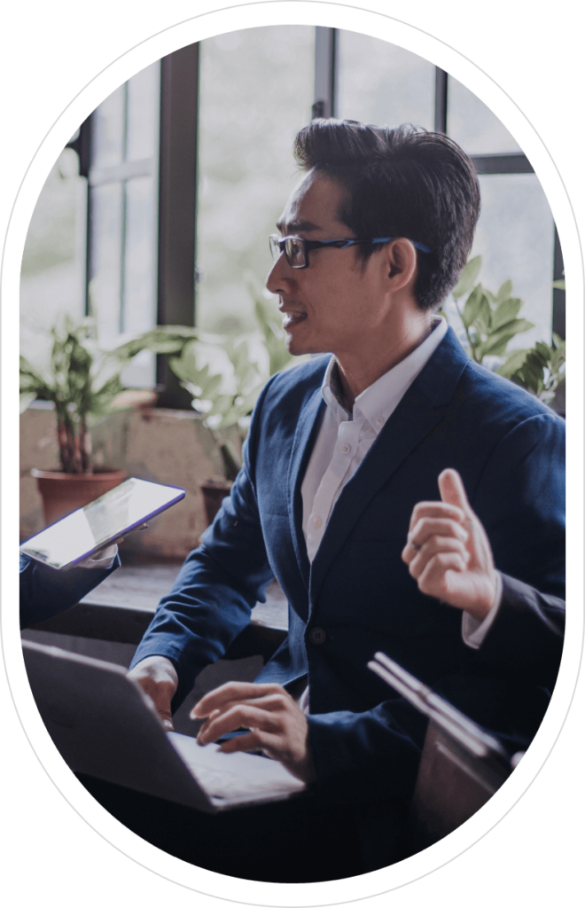 A man wearing glasses, a white shirt, and a blue blazer sitting at a desk with a laptop, engaged in a discussion with a person holding a tablet, in a room with large windows and green plants in the background.