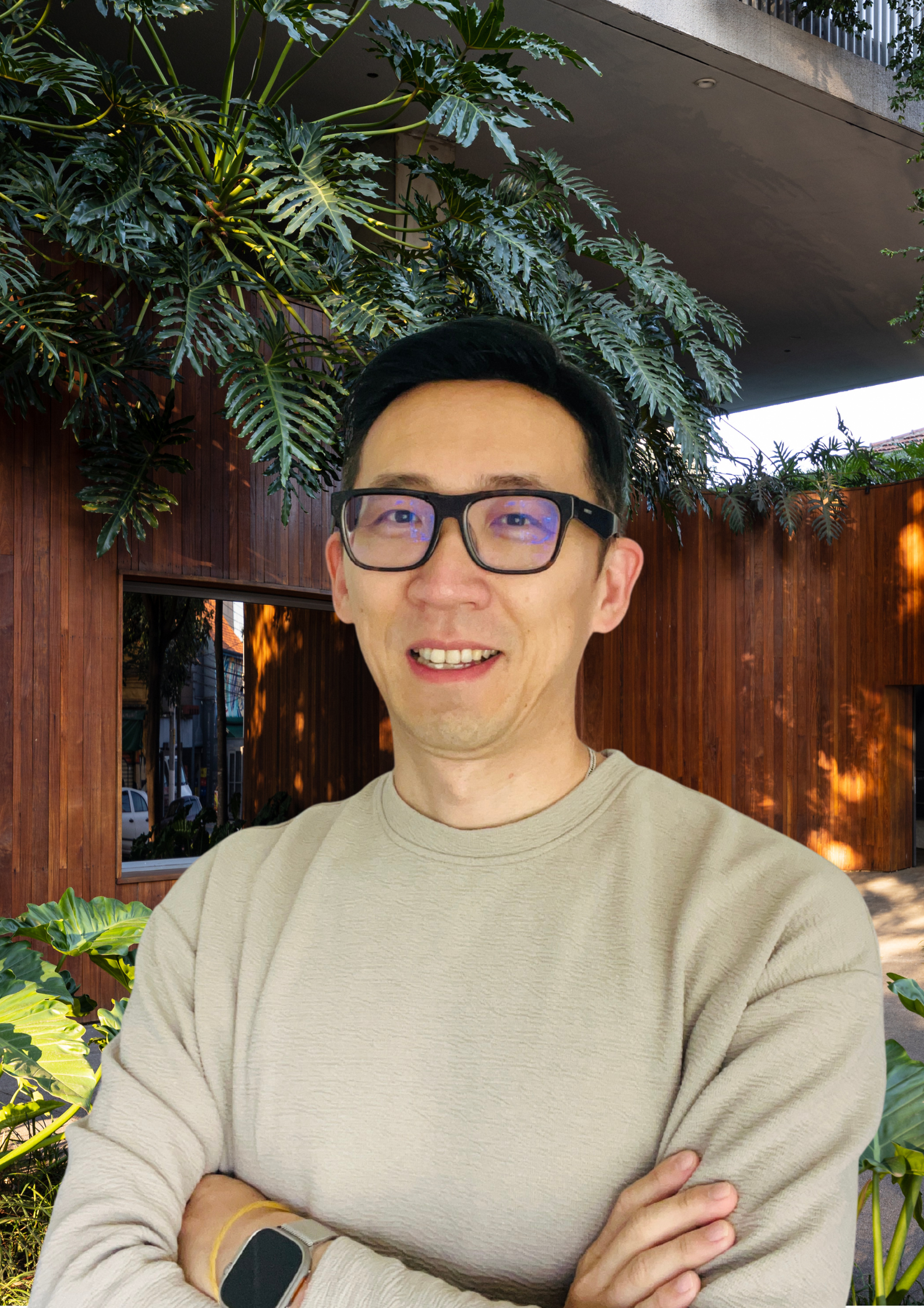 A man with black hair wearing glasses and a beige sweatshirt standing outdoors in front of a wooden building surrounded by green plants and trees.