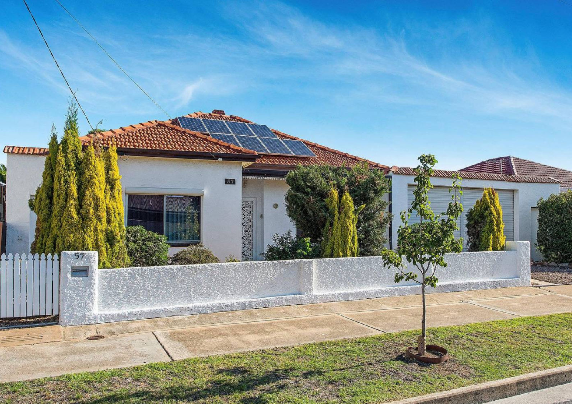 A white house with a red tiled roof, solar panels, and a small front yard featuring a white fence, trees, and shrubs, under a clear blue sky.