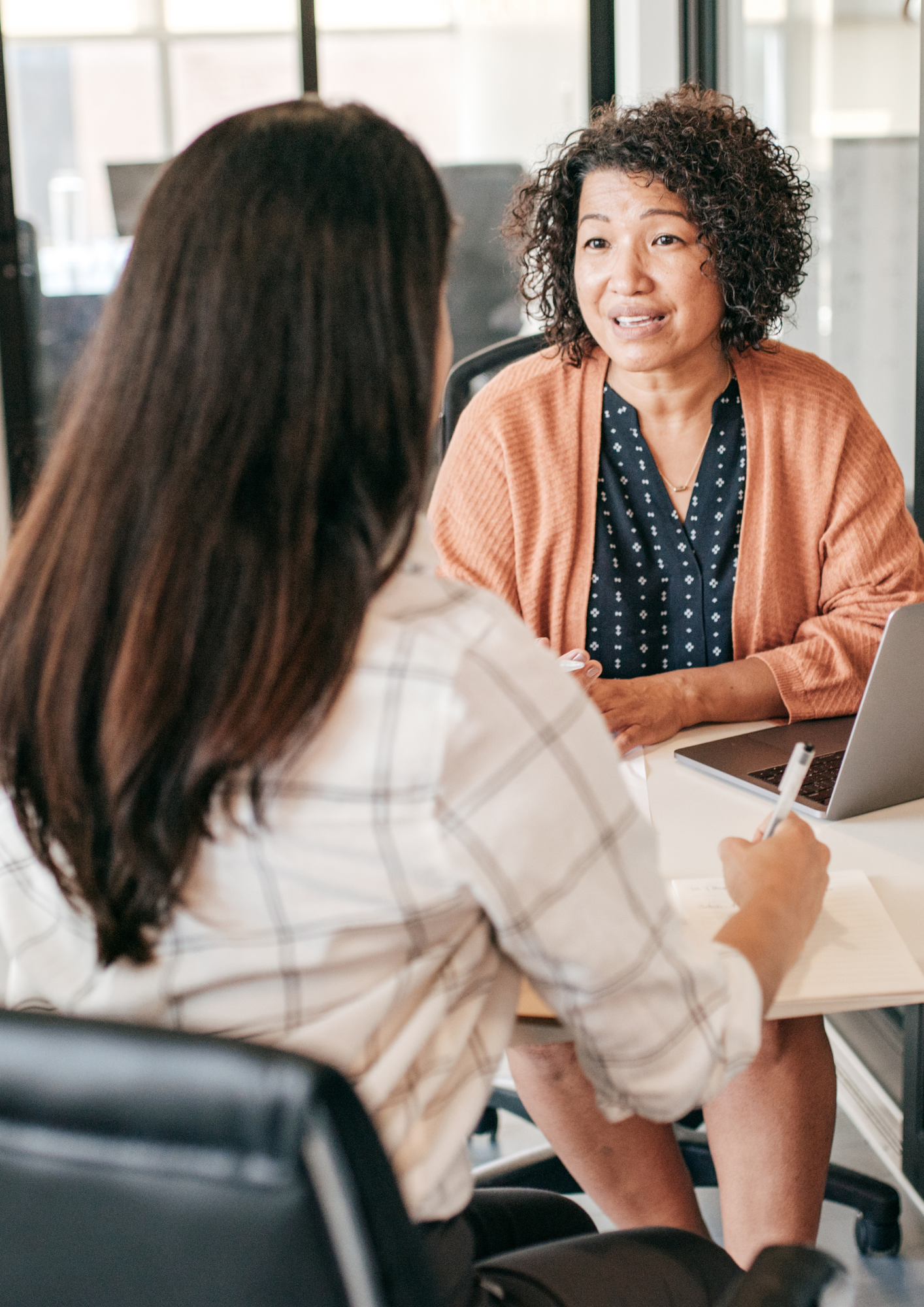 A woman with curly hair, wearing a rust-colored cardigan and a blue blouse, has a conversation with a woman with long dark hair, wearing a plaid shirt, in an office setting.