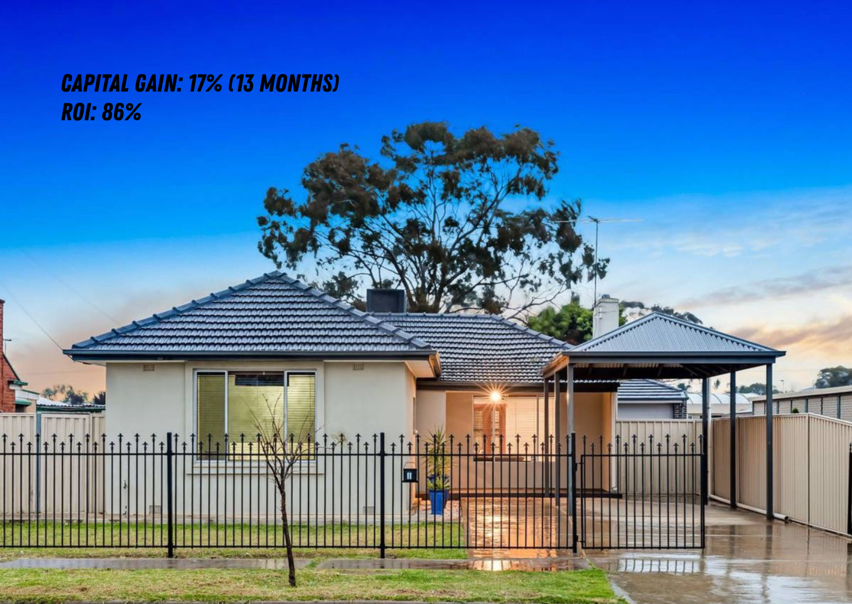 Front view of a house with a black metal fence, a small tree, and a wooden driveway gate. The house has a blue tiled roof, white exterior walls, and a covered porch. The sky is blue with a few clouds, and the setting sun is visible behind the house.