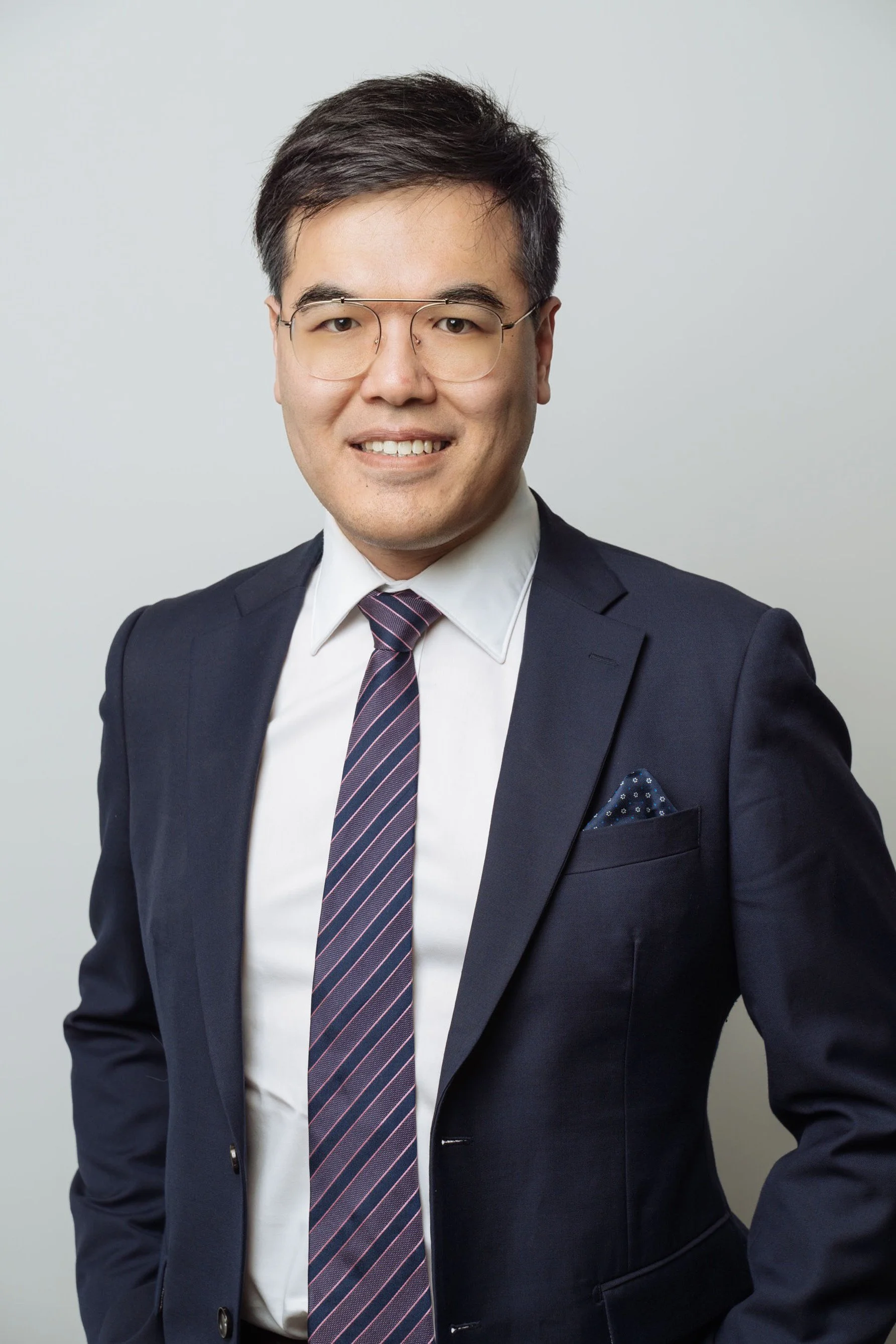 A man wearing glasses, a white shirt, a dark suit, and a tie, smiling in front of a plain light-colored background.