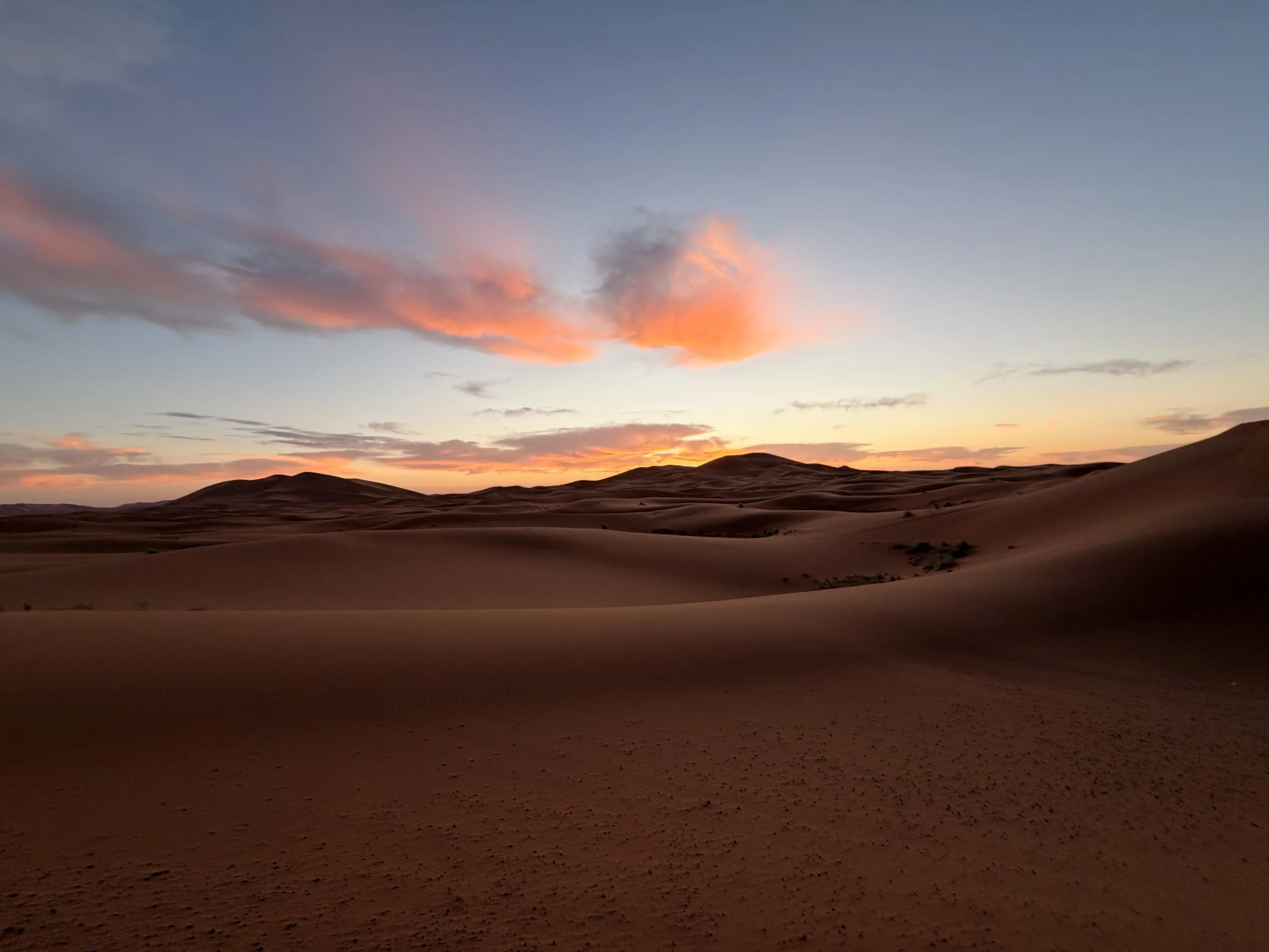 Sunset over the sand dunes of Merzouga in the Sahara Desert, Morocco, with orange and pink clouds stretching across the sky.
