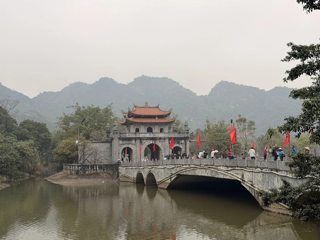 A traditional Asian-style building with curved roof and multiple levels, situated on a small island surrounded by water. A stone bridge with archways connects the island to the land, with people walking across it. Red flags line the bridge, and mountains are seen in the background, partially covered in fog or haze.
