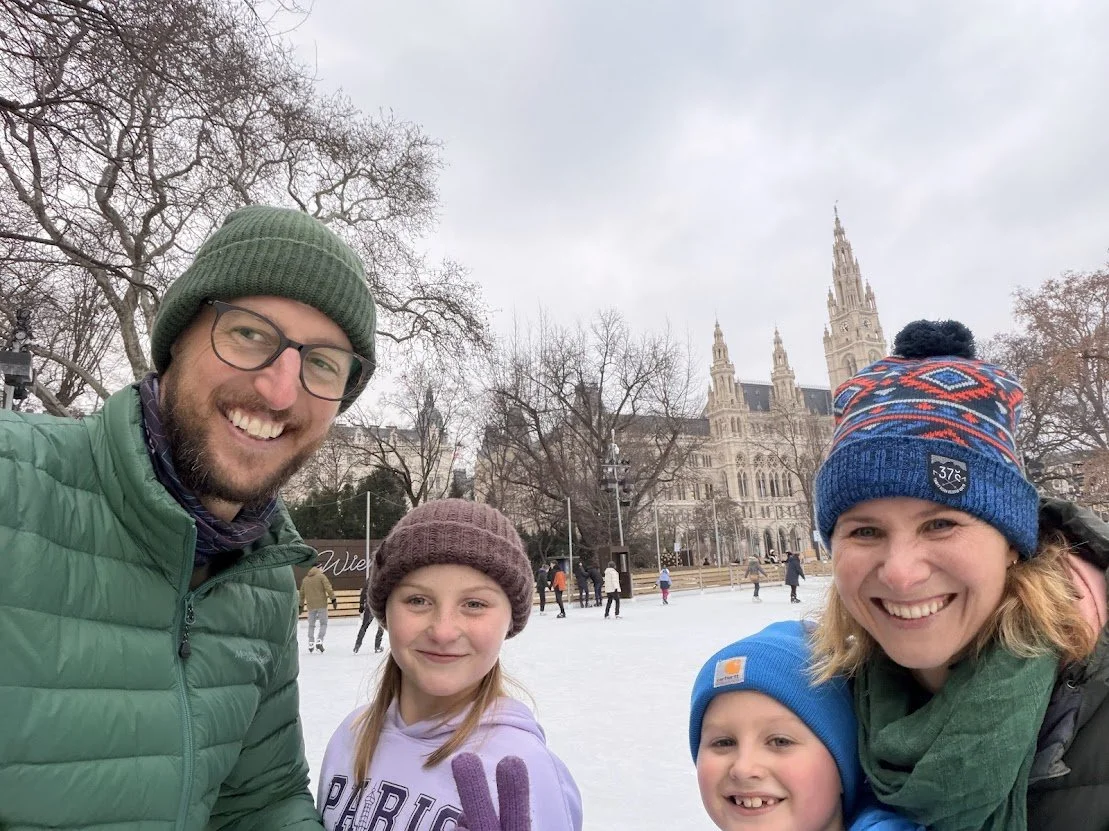 A family of four with two adults and two children enjoying ice skating outdoors in winter. They are dressed in warm clothing, with a historic building and leafless trees in the background.