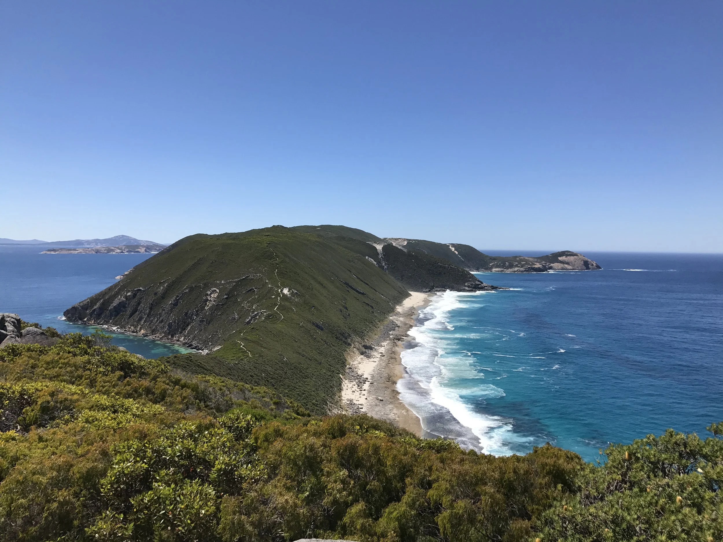 Coastal landscape with green hills, sandy beach, and ocean waves under a clear blue sky.