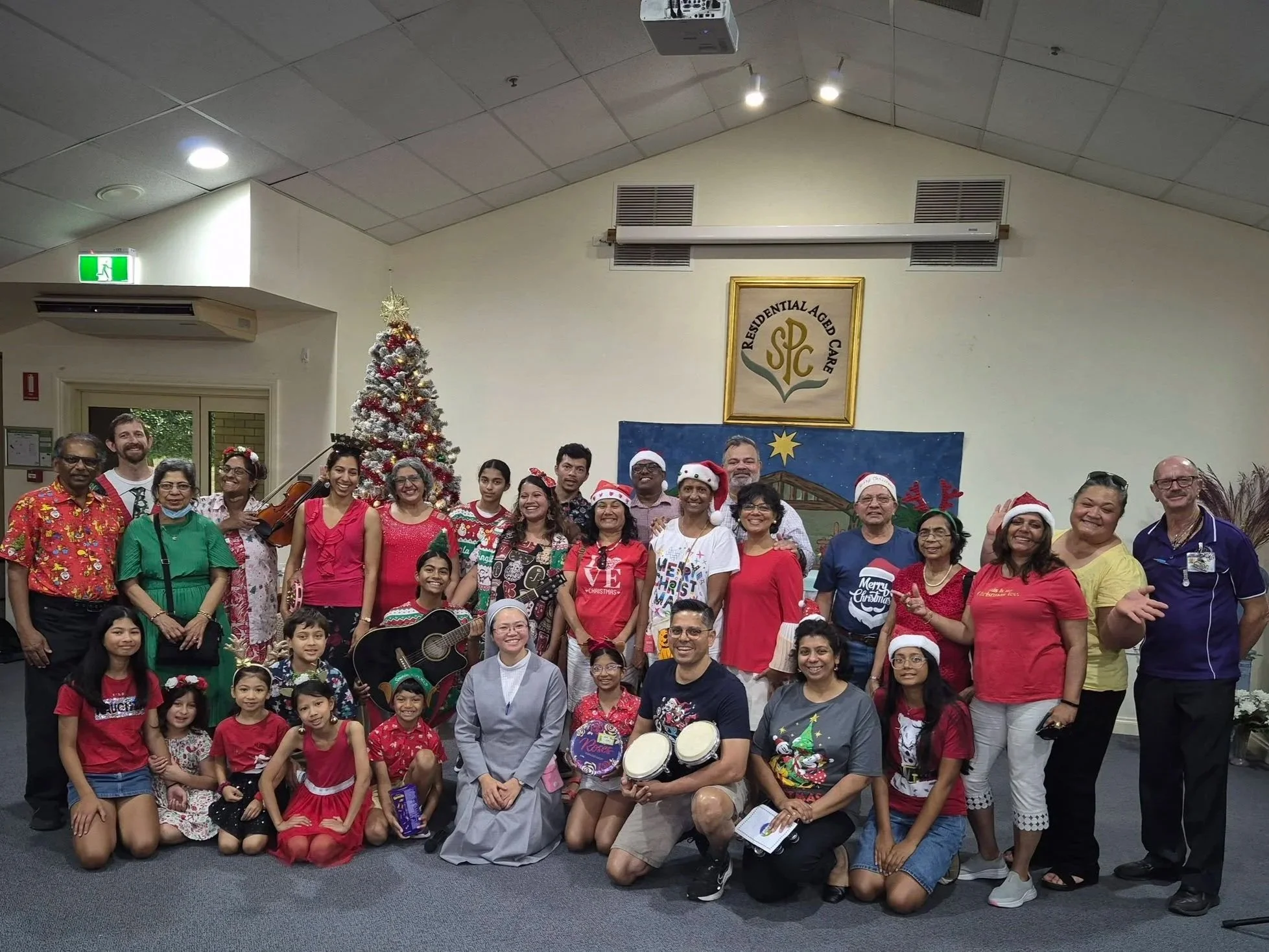 A large group of people celebrating Christmas indoors, some wearing Santa hats and reindeer headbands, with a decorated Christmas tree in the background. The group includes children and adults, some holding musical instruments, and they are all smili