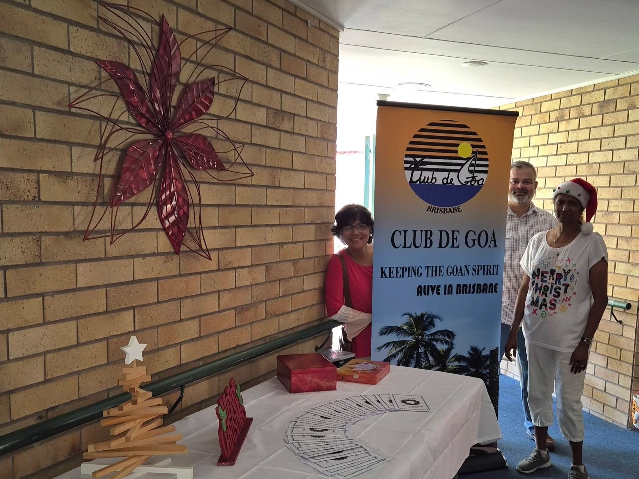 Three people standing near a table at a holiday event. One woman is wearing a Santa hat, and there is a Christmas tree and holiday decorations on the table, including a small wooden Christmas tree with a star on top, and a sign that reads 'Merry Chri