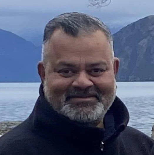 Close-up portrait of a man with short dark hair and a beard, wearing a dark jacket, outdoors near a body of water with mountains in the background.
