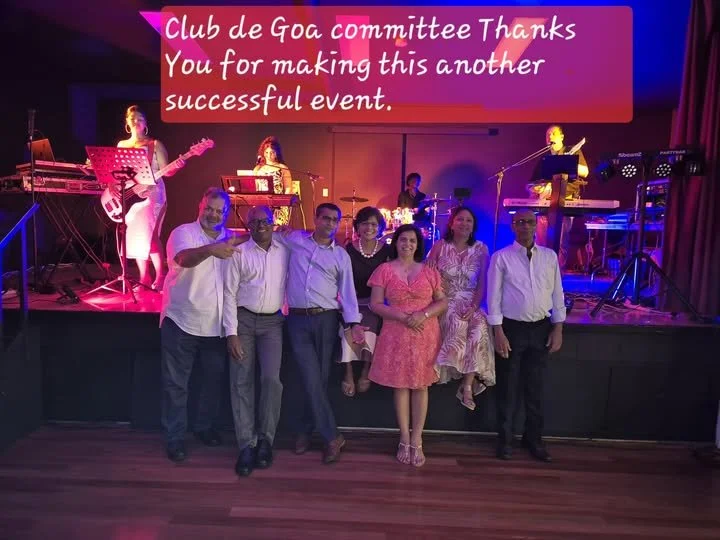 Group of seven people, five men and two women, standing in front of a stage with a live band performing. The stage has colorful lighting and musical instruments. A pink and purple banner on the stage reads, 'Club de Goa committee Thanks You for makin