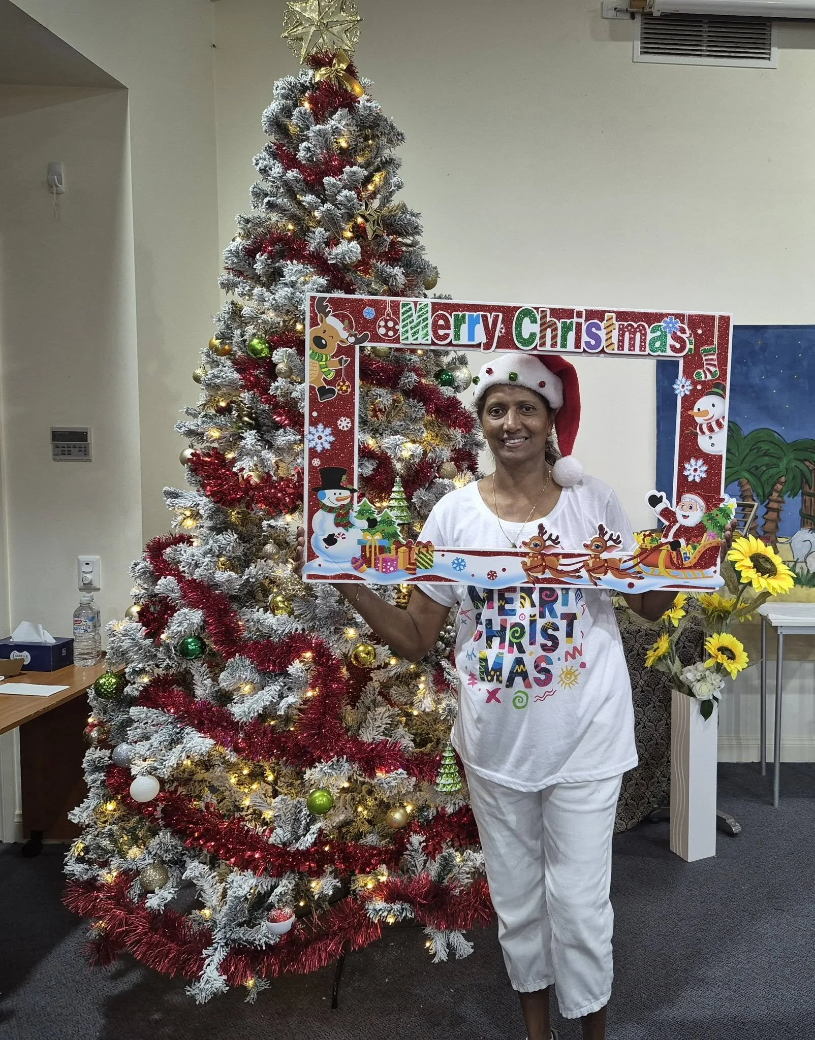 A woman wearing a Santa hat and a colorful Christmas-themed T-shirt stands in front of a decorated Christmas tree, holding a festive picture frame with Christmas decorations and the words "Merry Christmas" at the top.