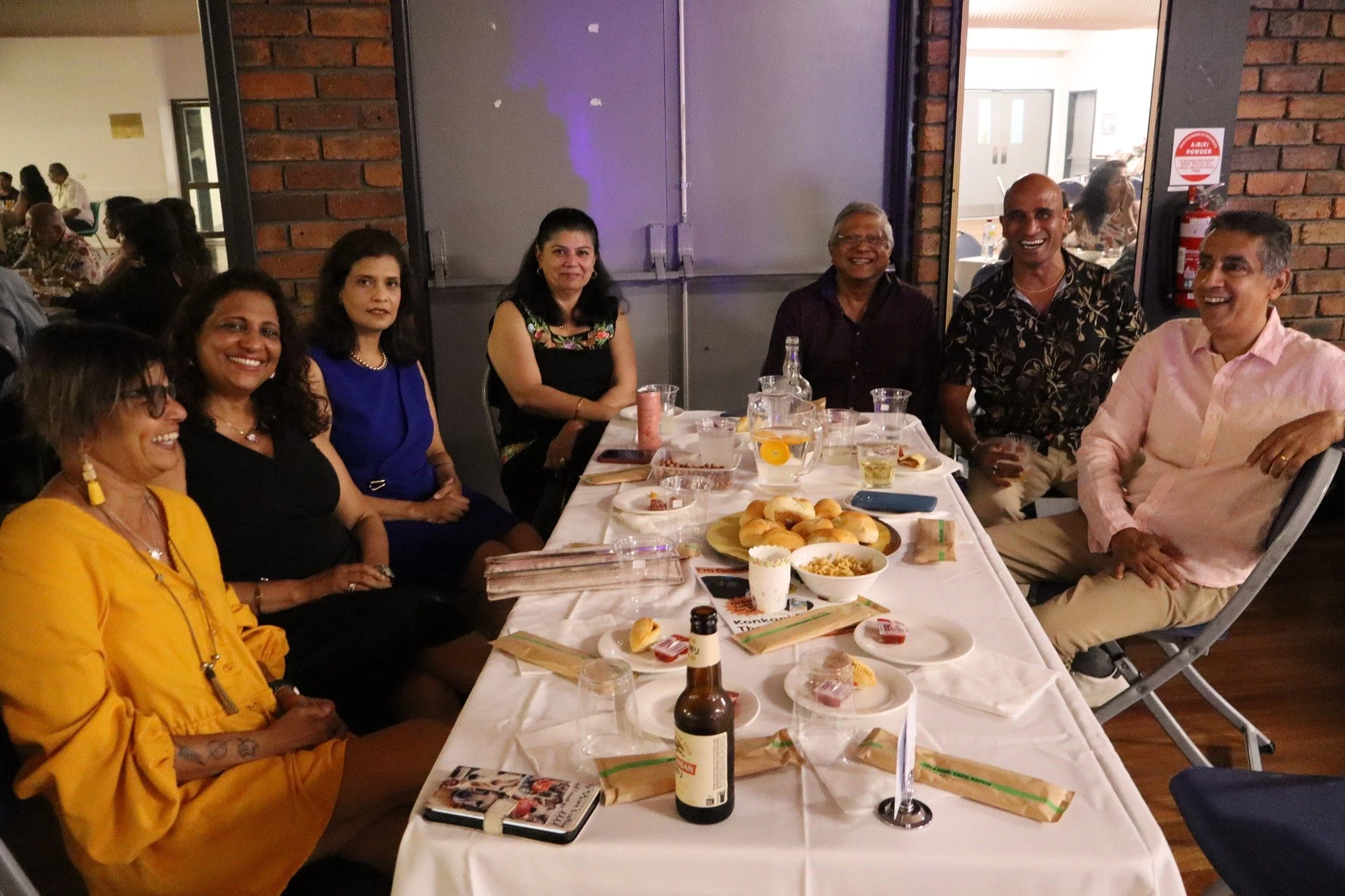 A group of eight people sitting around a table at a social gathering, smiling and enjoying food and drinks, in an indoor restaurant or event space.