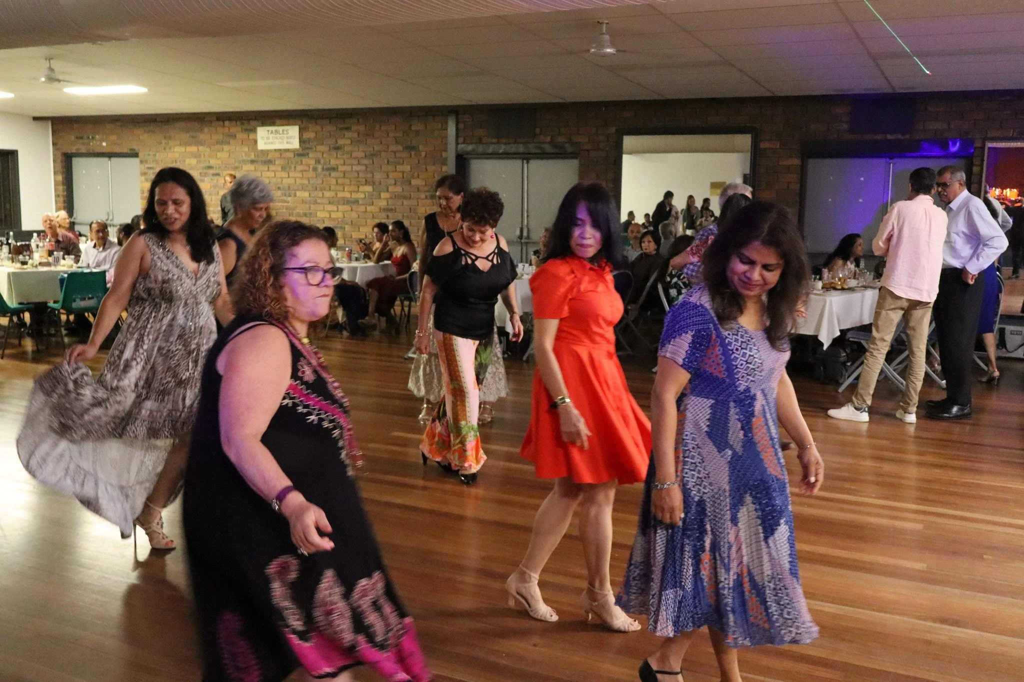 Group of women dancing on a wooden floor at a social event in a hall with brick walls and tables with people dining in the background.