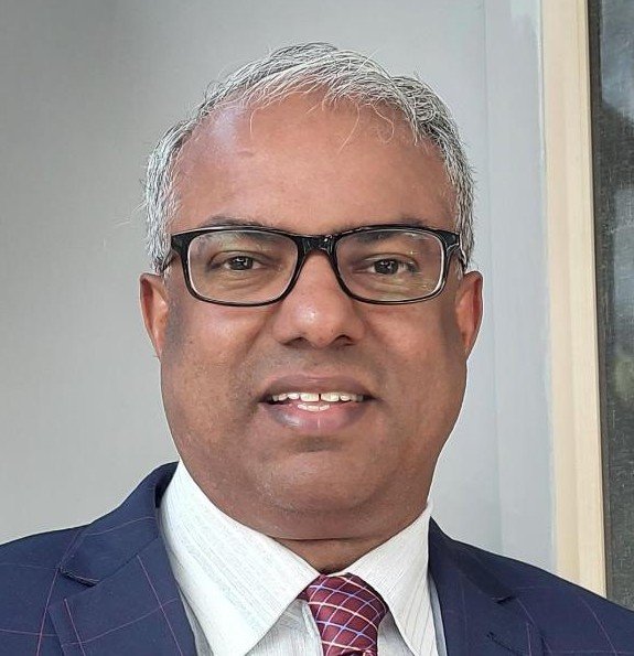 A man with gray hair and glasses wearing a white shirt, a patterned tie, and a dark suit standing indoors near a window.