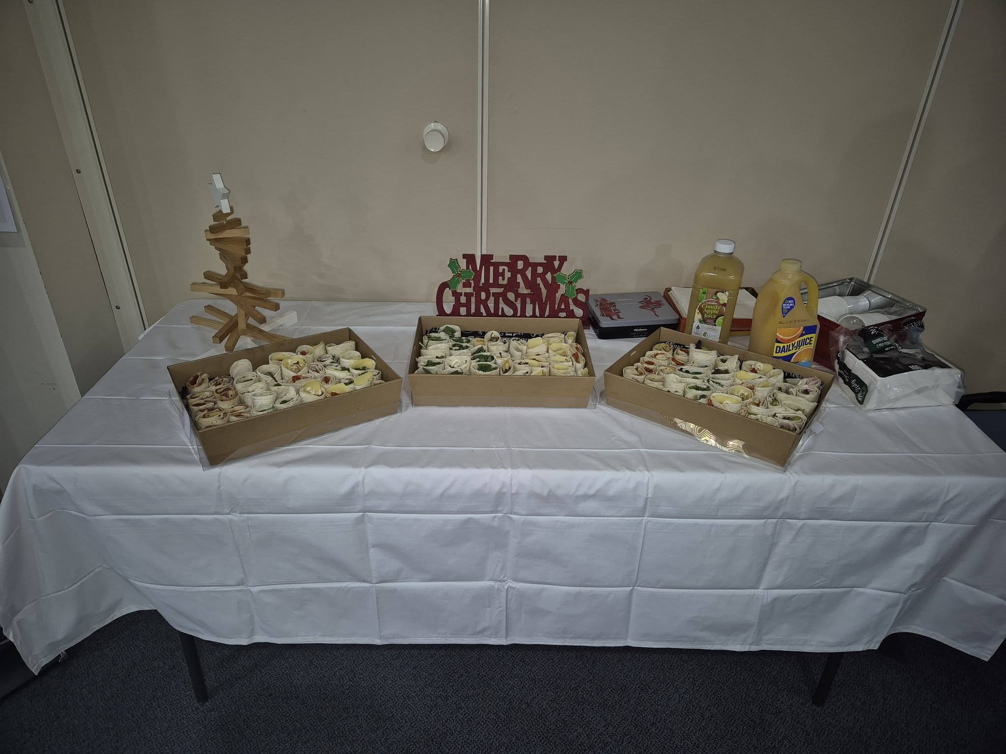 Table with three boxes of assorted rolled sandwiches, a Christmas sign, a wooden Christmas tree, juice bottles, snack bars, and holiday decorations.
