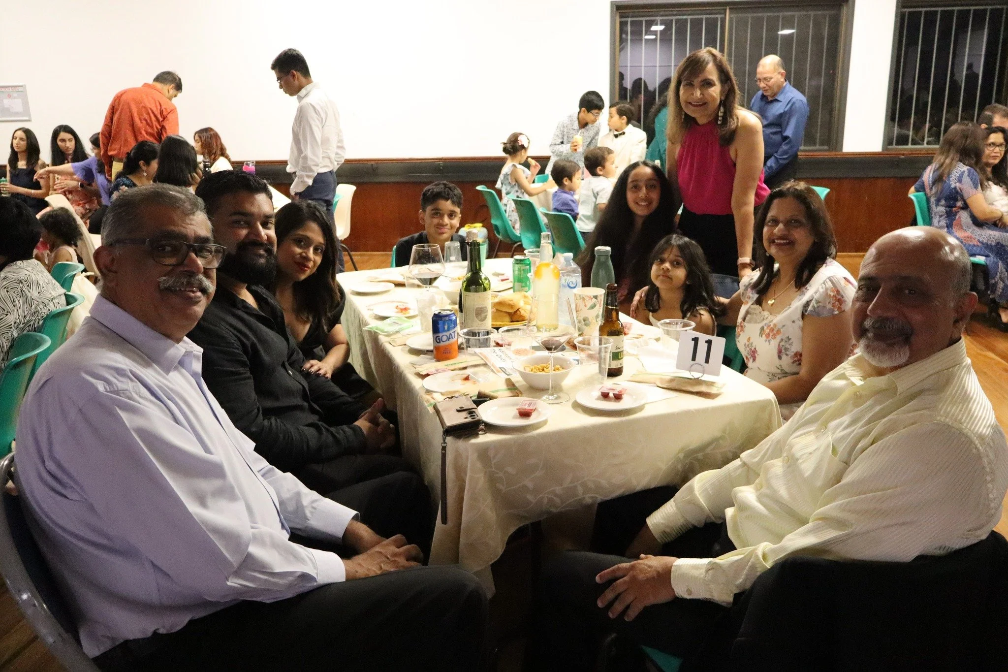 A group of nine people, including adults and children, sitting around a banquet table at a social gathering or celebration, with food and drinks on the table, smiling for the camera in a large hall filled with other guests.